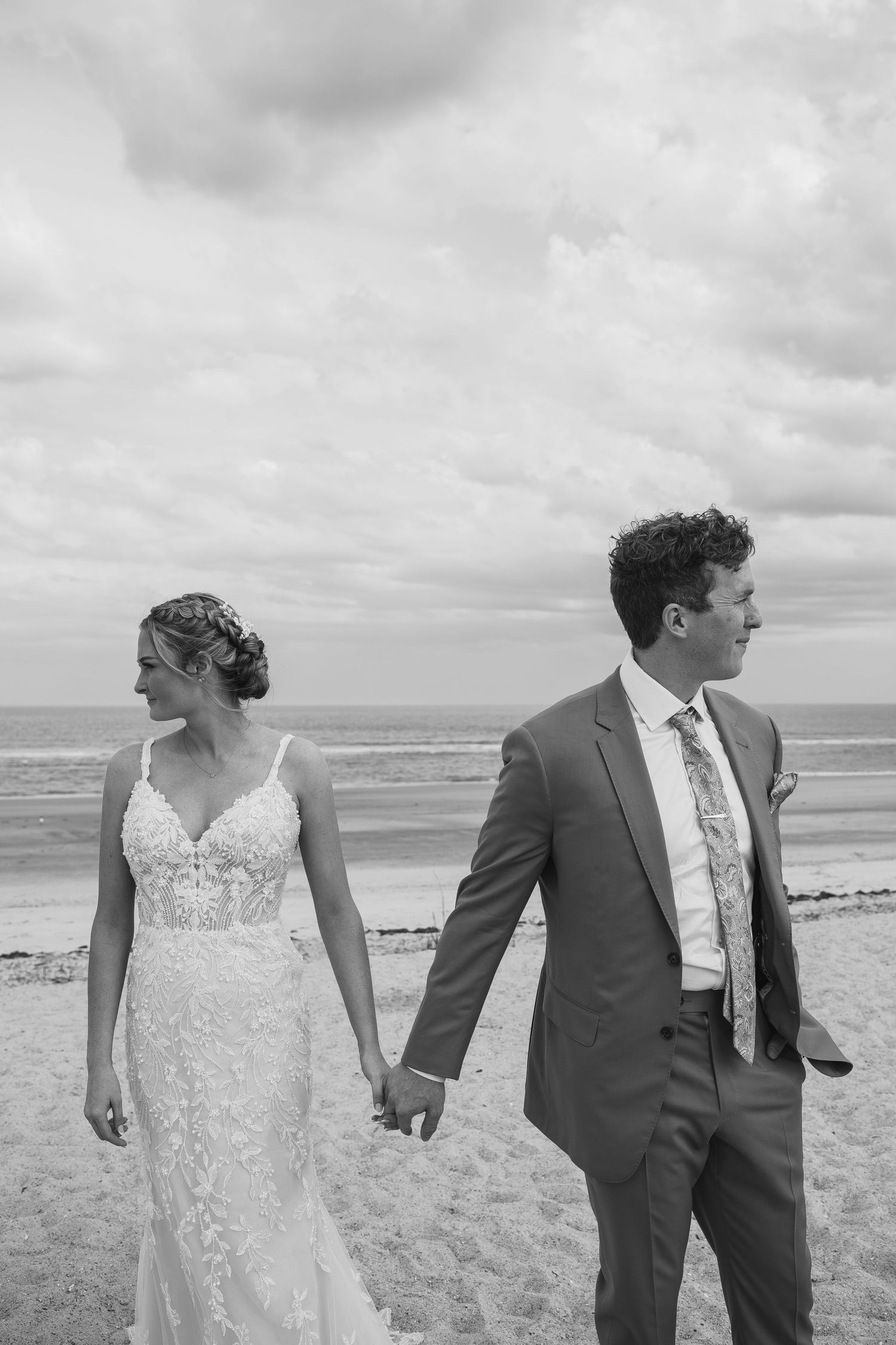 Black and white photo of a bride and groom holding hands on a beach, with ocean waves and cloudy skies behind them.
