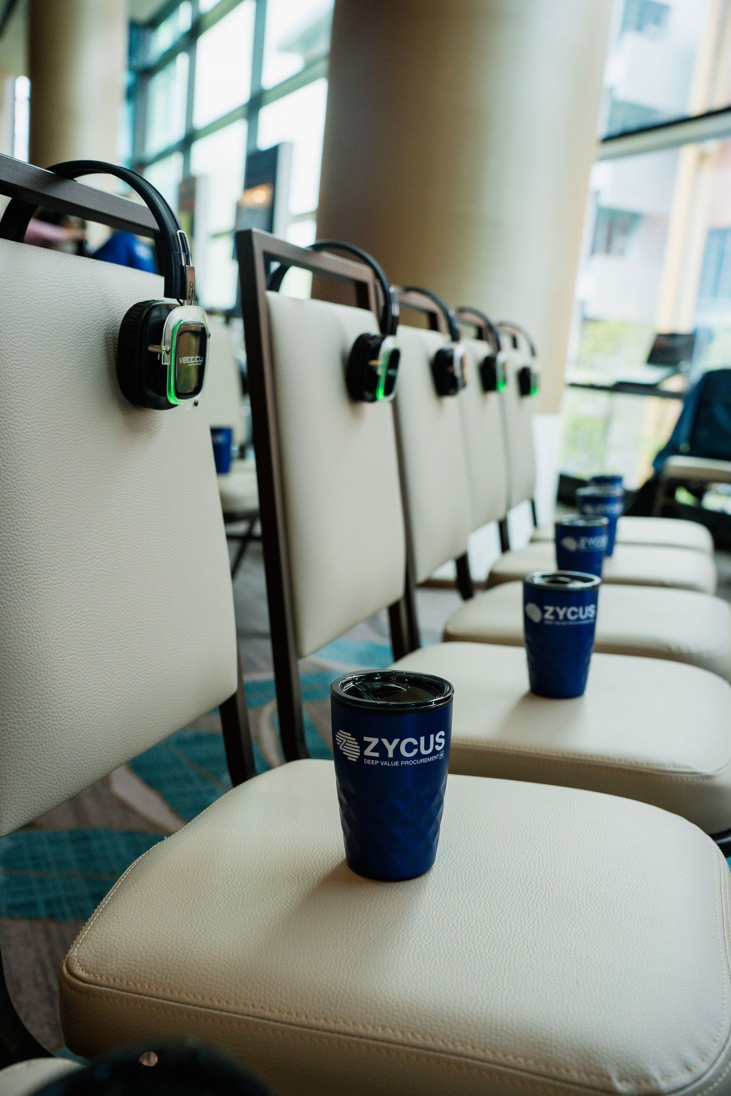 Zycus branded blue tumblers and headsets placed on cream chairs at a corporate conference event.