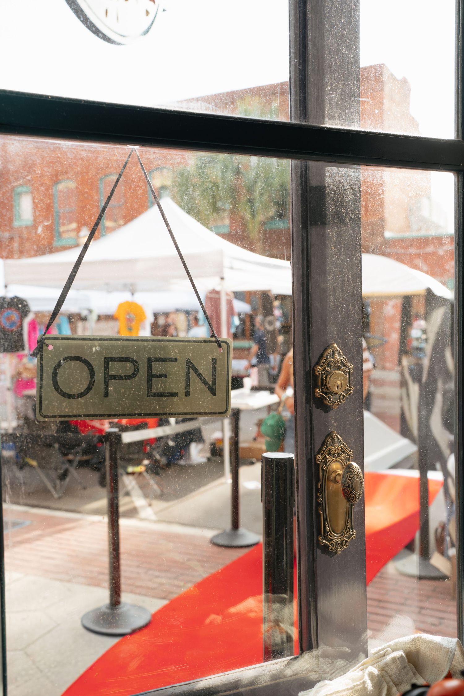 A vintage brass Open sign hangs on a glass door with ornate gold hardware, overlooking a busy outdoor market.