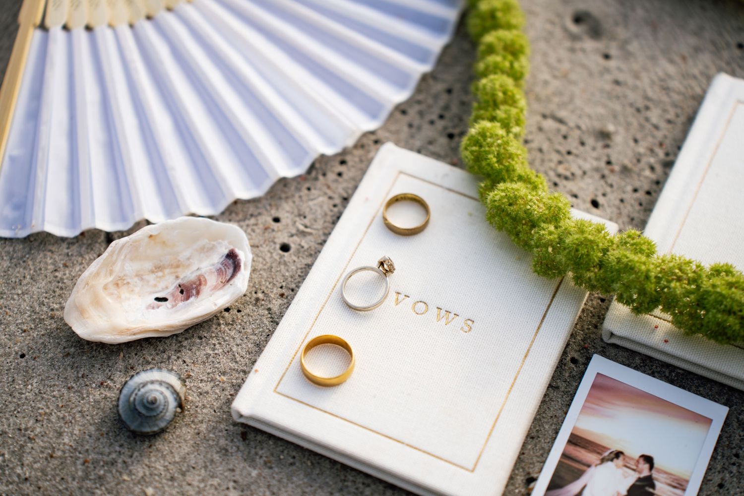 Wedding rings rest on a white paper holder decorated with green moss against a textured gray background.