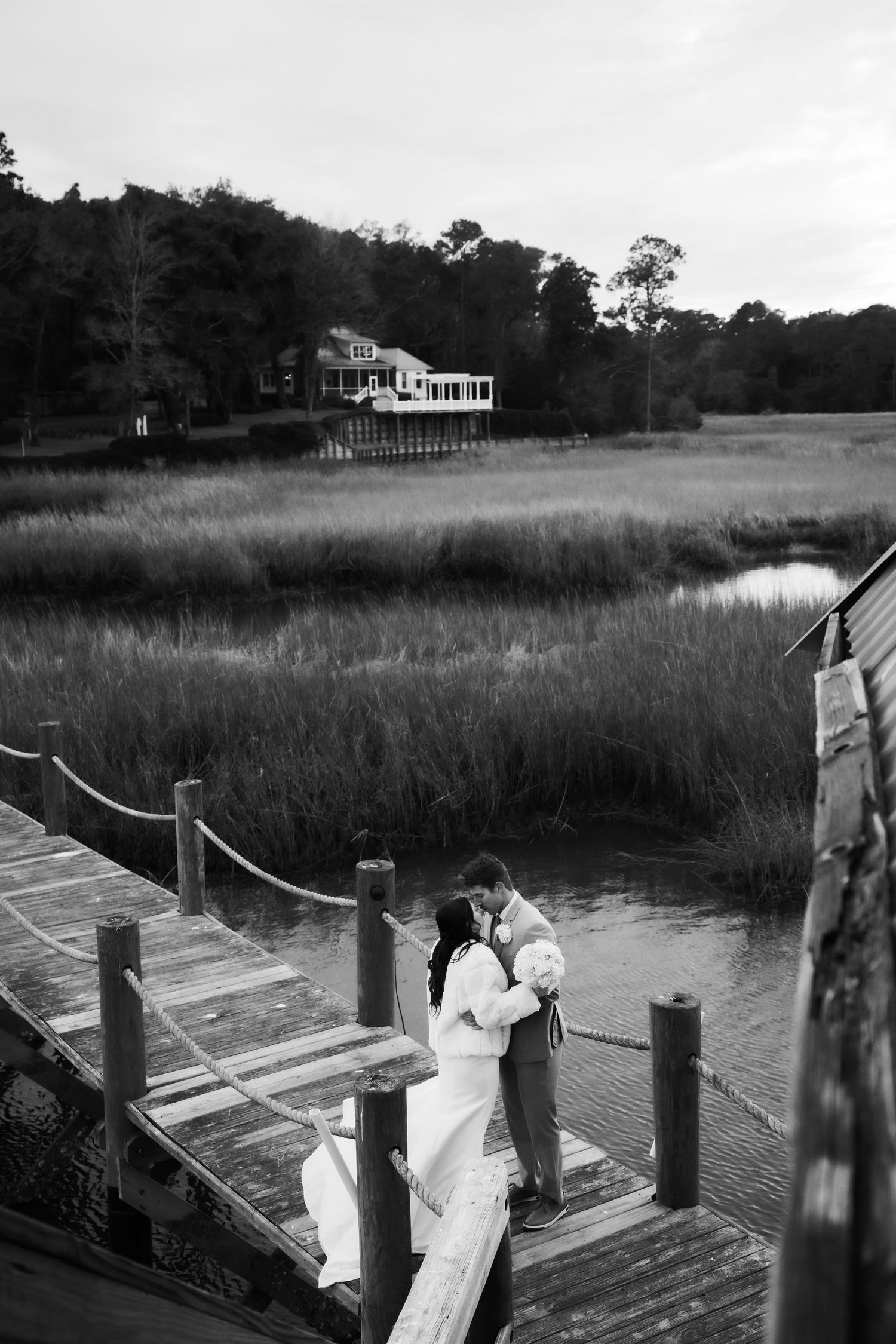 A couple embraces on a wooden dock overlooking a scenic marsh landscape with waterfront homes in the background.