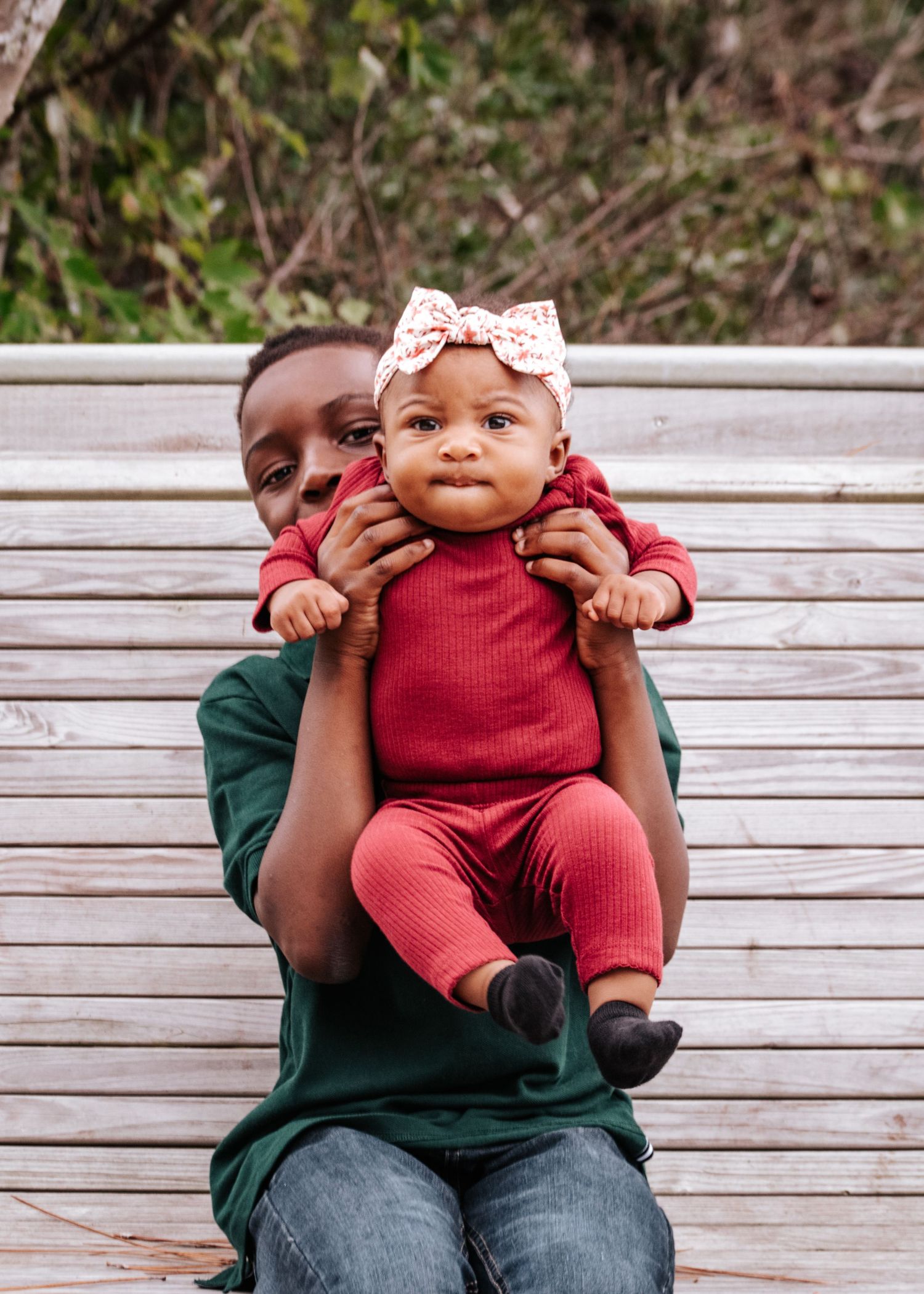 Loved one holds a baby wearing a pink bow headband and red outfit on wooden bench.