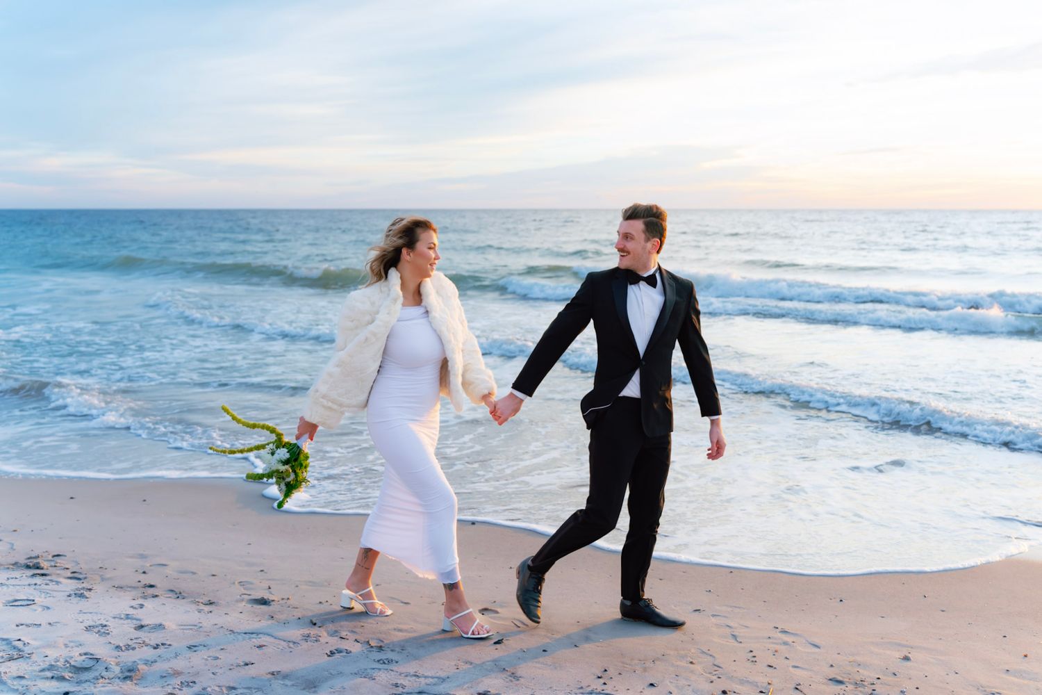 A couple in wedding attire walks along the shoreline at sunrise holding hands and bouquet on a sandy beach in winter on Amelia Island