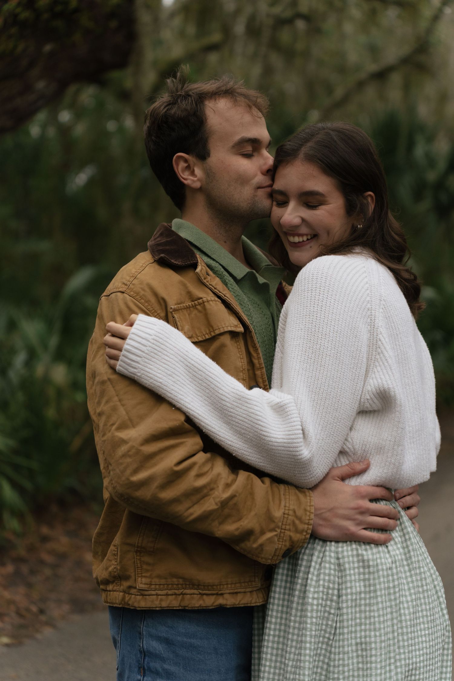 A couple in casual attire share intimate moments in a wooded outdoor setting, wearing brown jacket and white sweater.