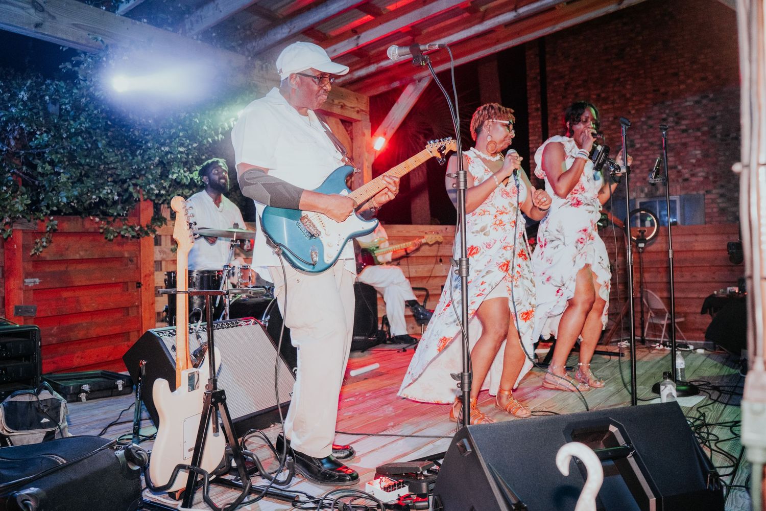 Live band performing outdoors, guitarist in white hat and two female singers in floral dresses on a rustic wooden stage.