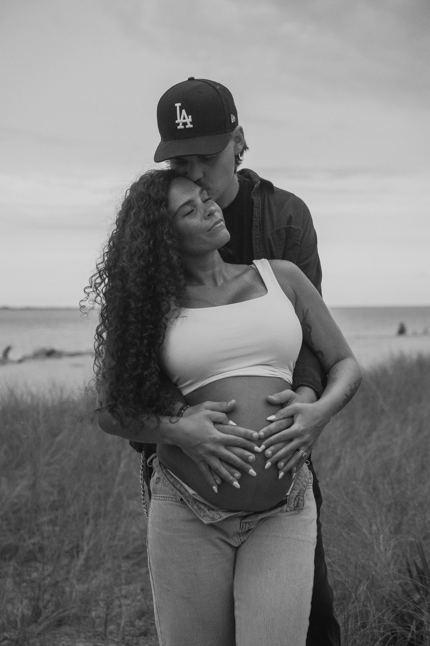 Intimate black and white maternity photo of couple embracing on grassy hill.