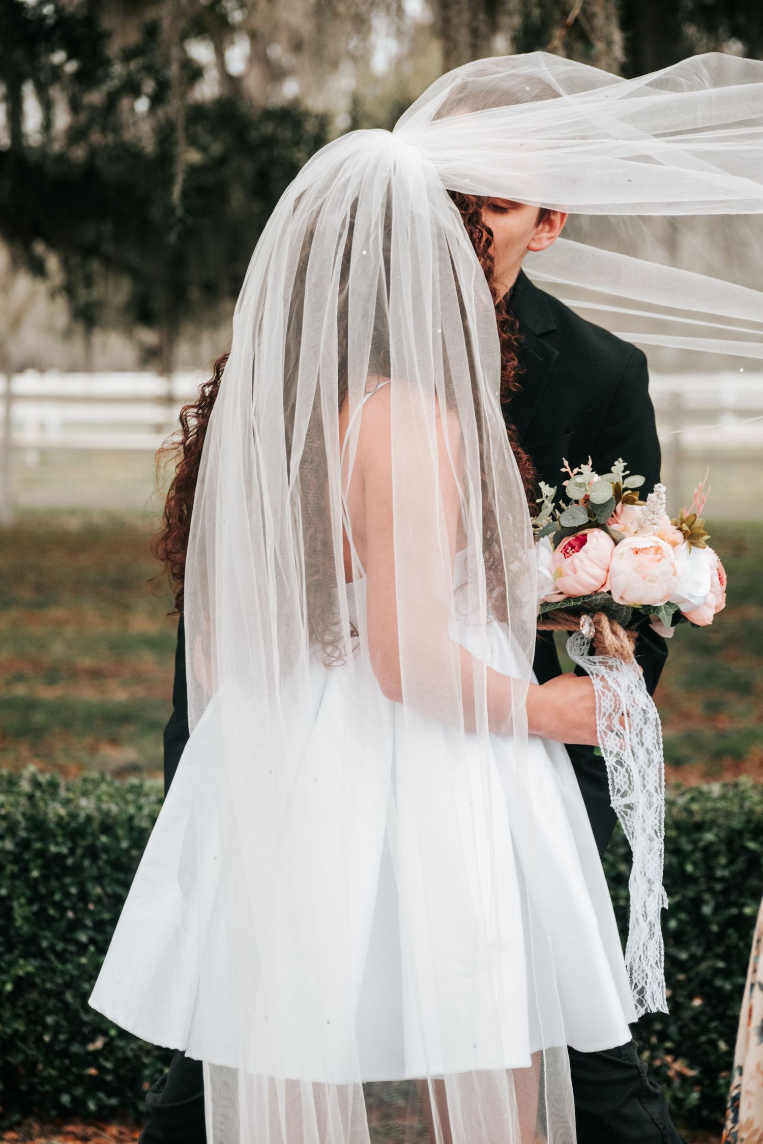 A romantic wedding moment captured as a couple embraces under a flowing white veil, with pink roses in hand.