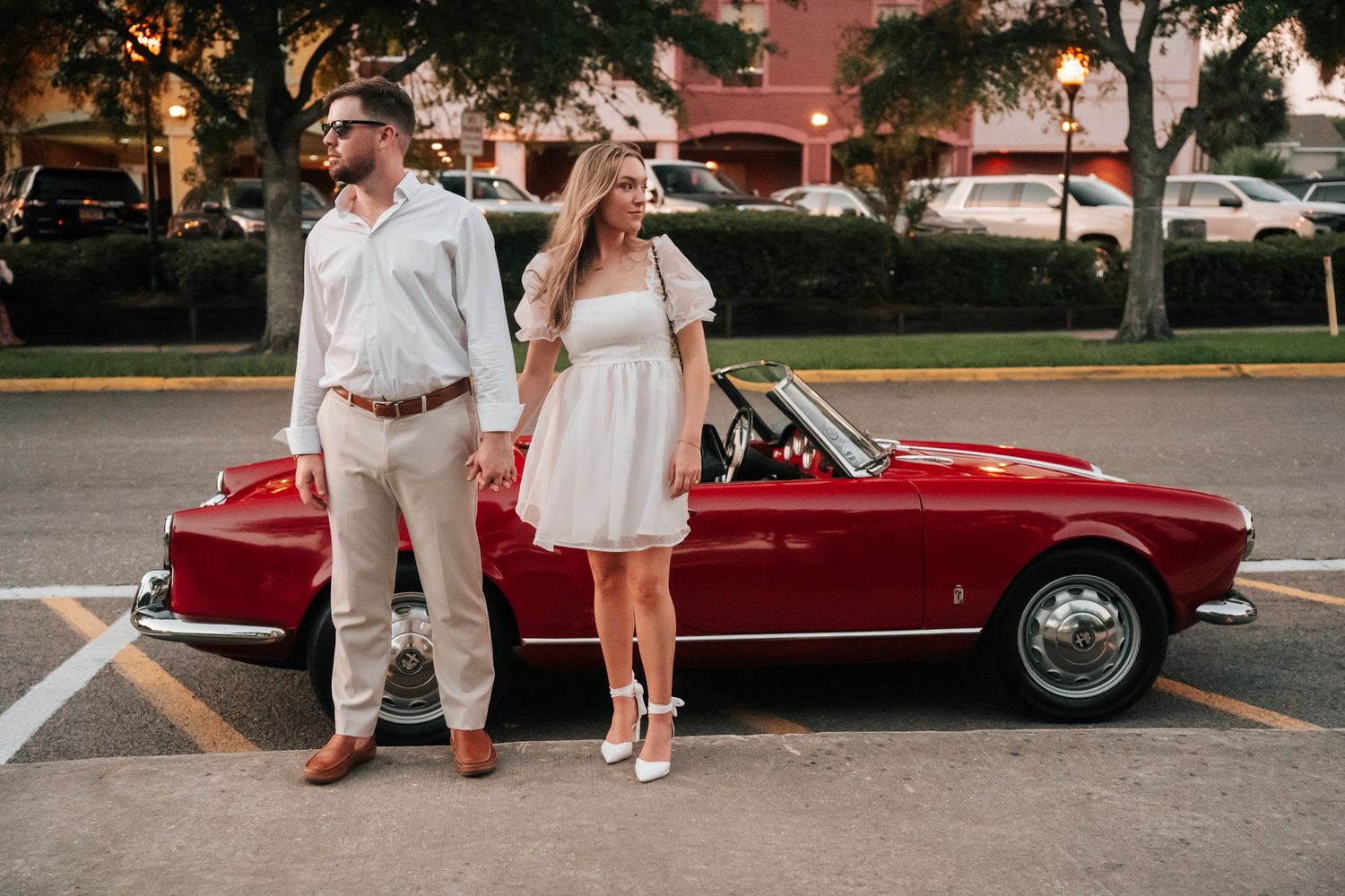 Stylishly dressed couple poses beside a classic red vintage car in a parking lot at sunset.