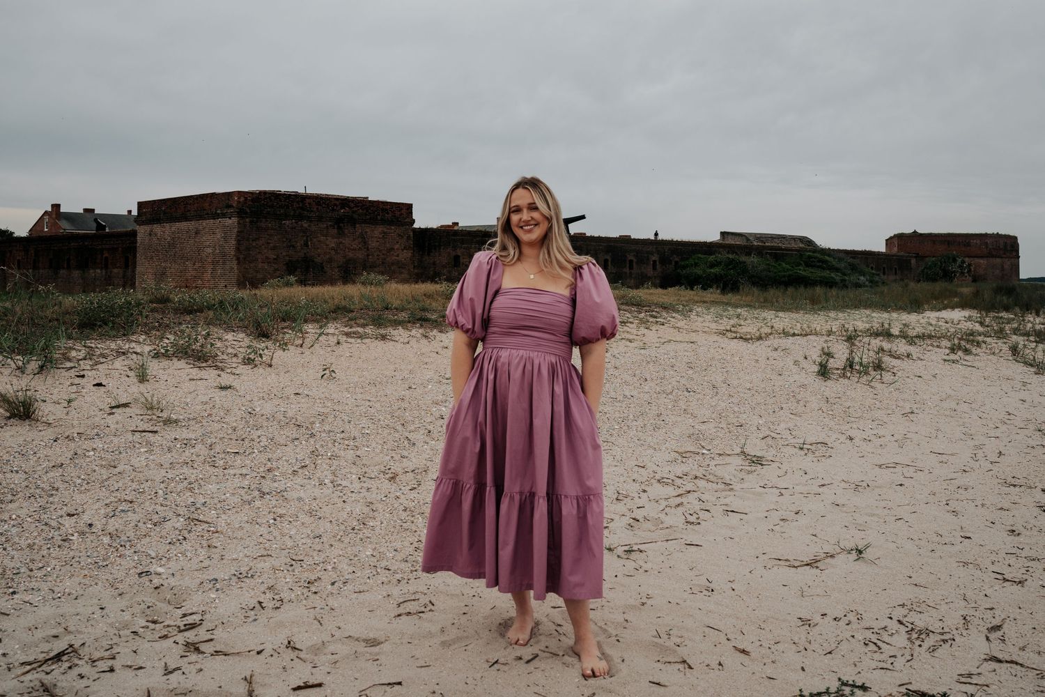 A person in a mauve dress stands barefoot on sandy beach with old fort walls visible in the background. Fort Clinch State Park
