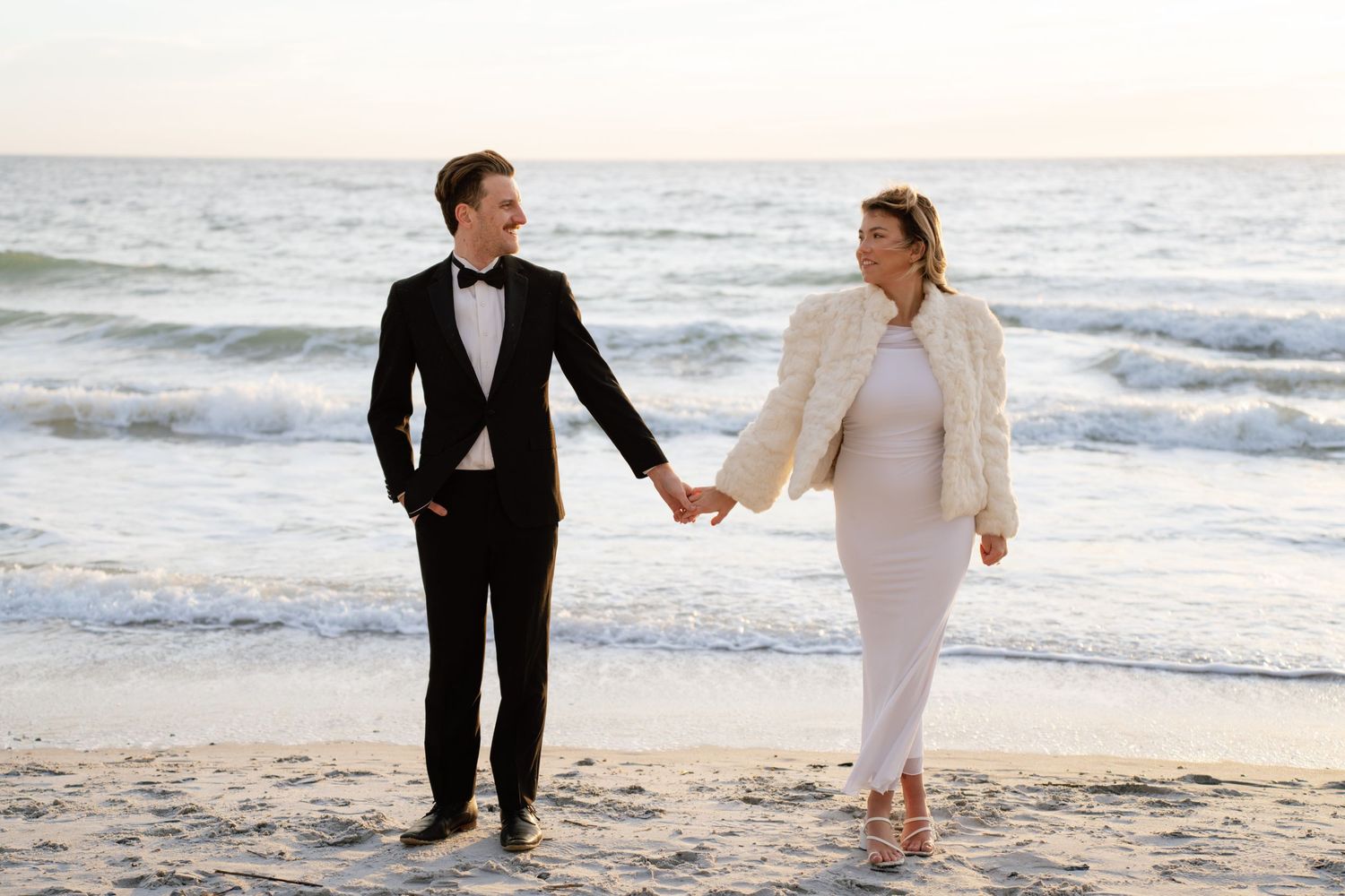 A couple in wedding attire walks hand in hand along a scenic beach at sunset with waves crashing in the background.