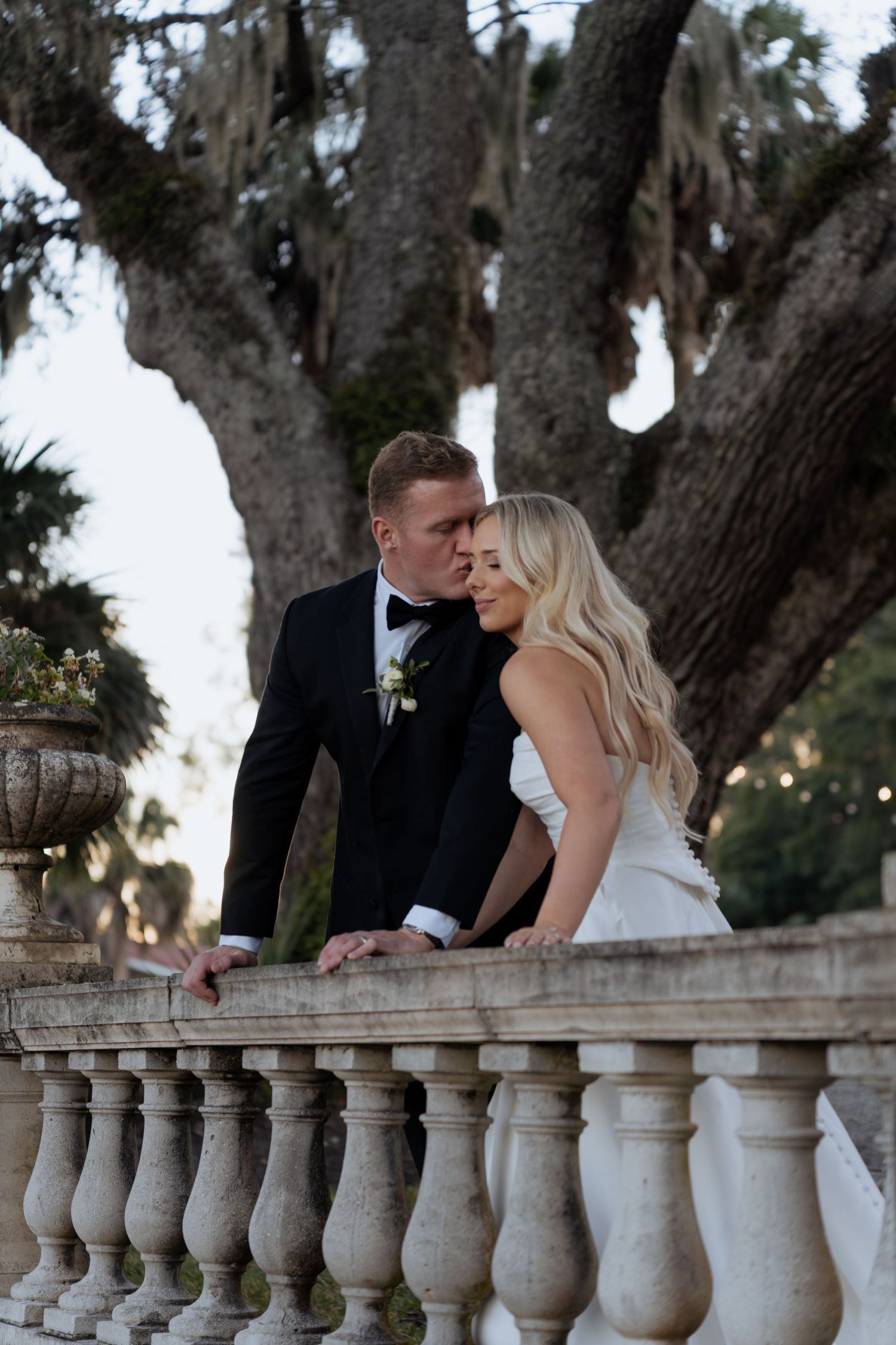 A romantic couple in wedding attire share intimate moments along a stone balustrade at sunset.