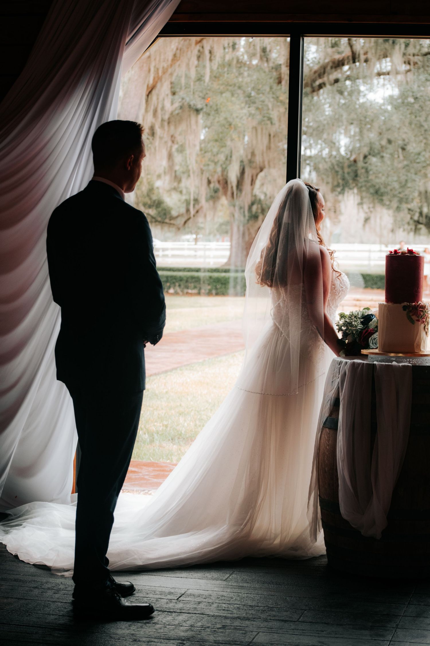 Silhouetted figures stand by a window in a series of romantic wedding photos with soft natural lighting and flowing veil.