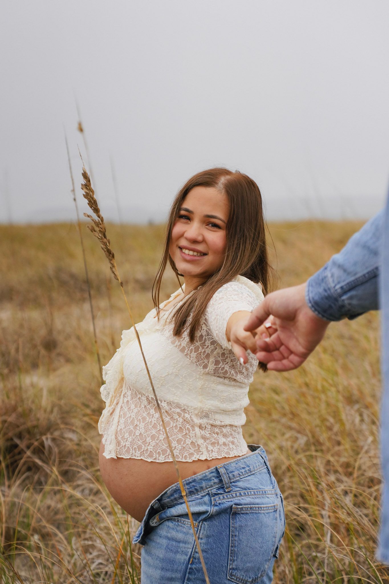 Person wearing a white lace top and jeans stands in a golden field of dry grass at sunset.