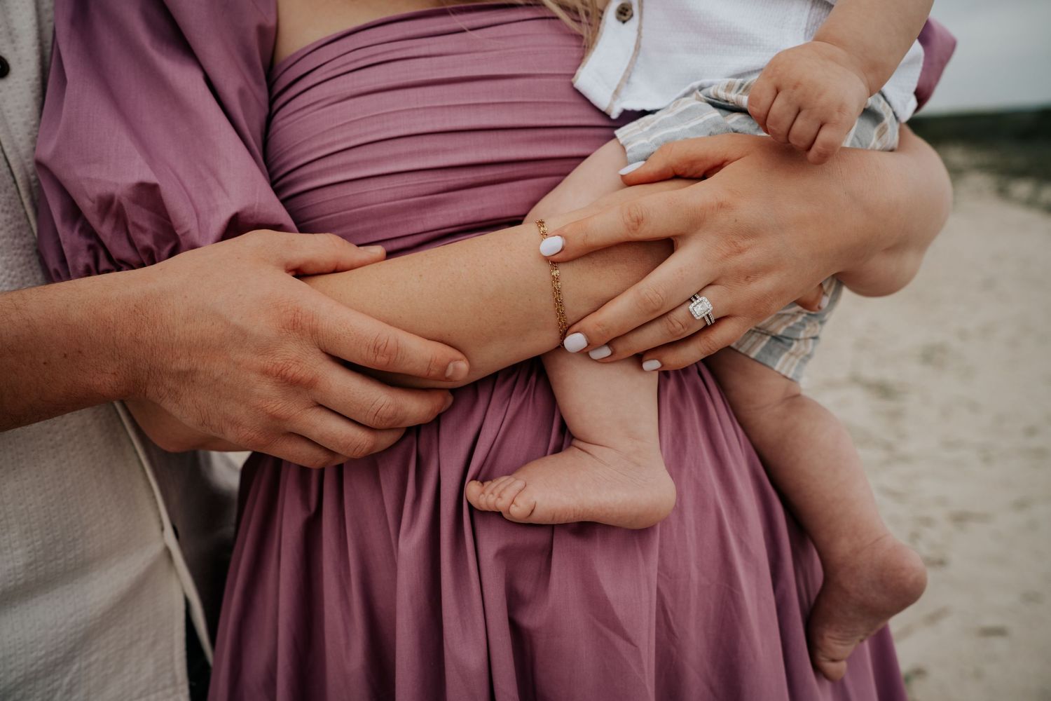 Close up of hands holding a baby while wearing a mauve colored dress outdoors.