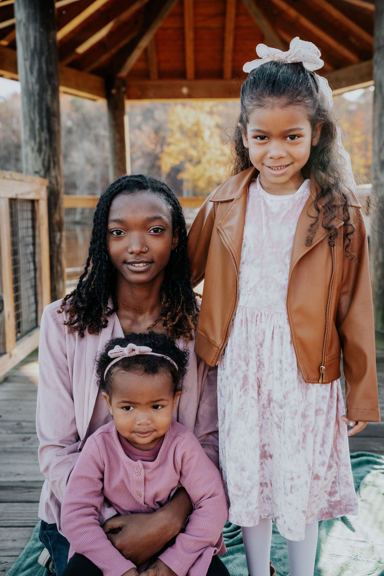 Family of three in autumn colors posing under wooden gazebo.