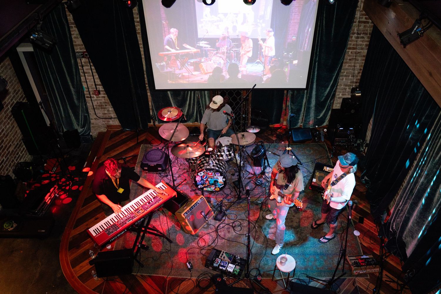 Overhead view of a live band performing on stage with keyboards, drums, and guitars under red stage lighting.