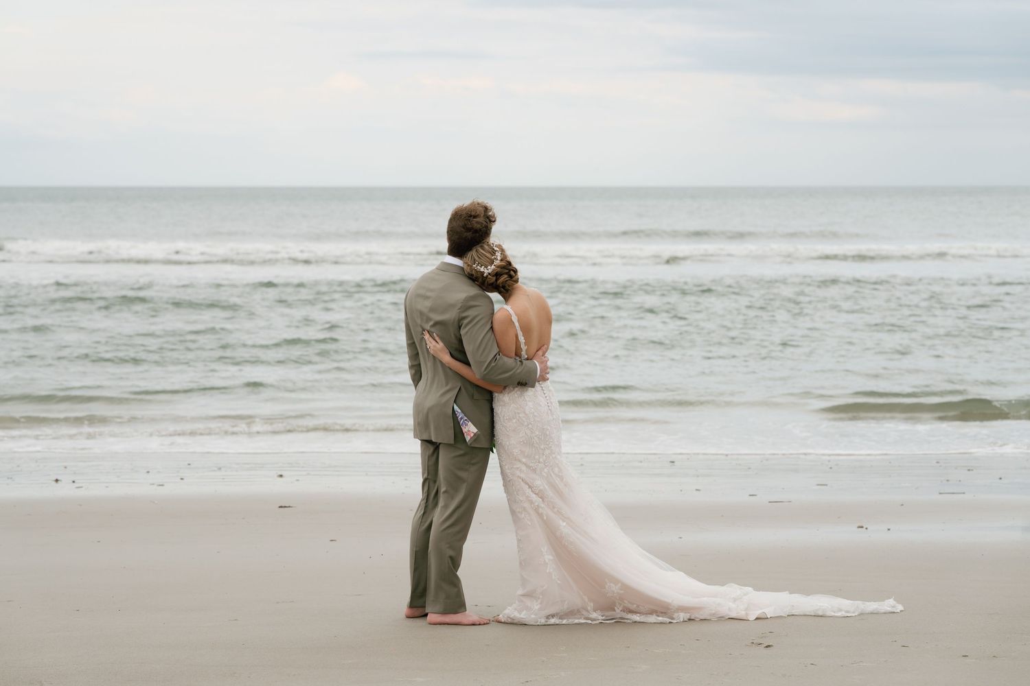 Bride and groom embrace on a sandy beach with ocean waves behind them during their romantic seaside wedding ceremony.