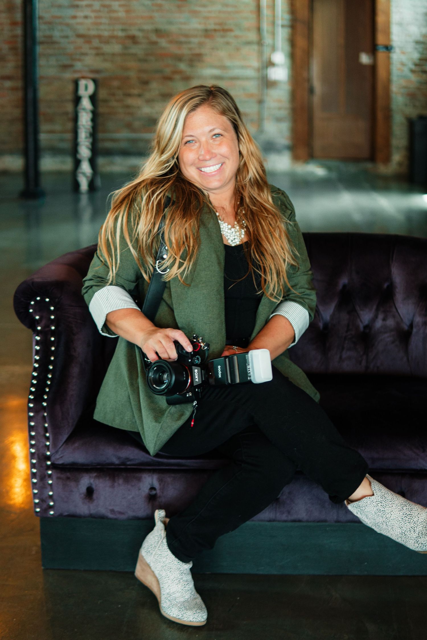 Professional photographer sitting on dark velvet couch holds camera while wearing green blazer and white sneakers.