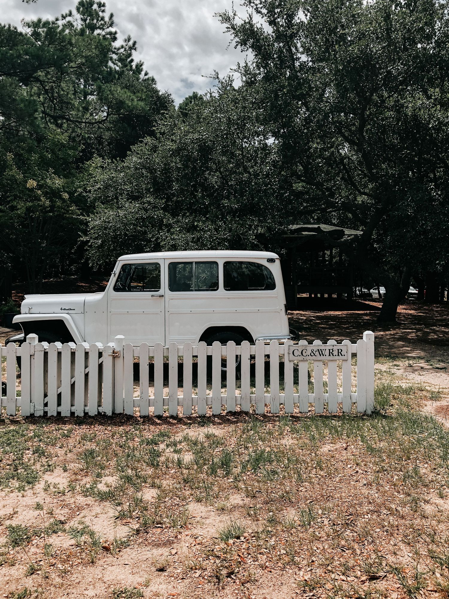 Vintage white Willys station wagon parked behind a white picket fence with lush green trees in the background.