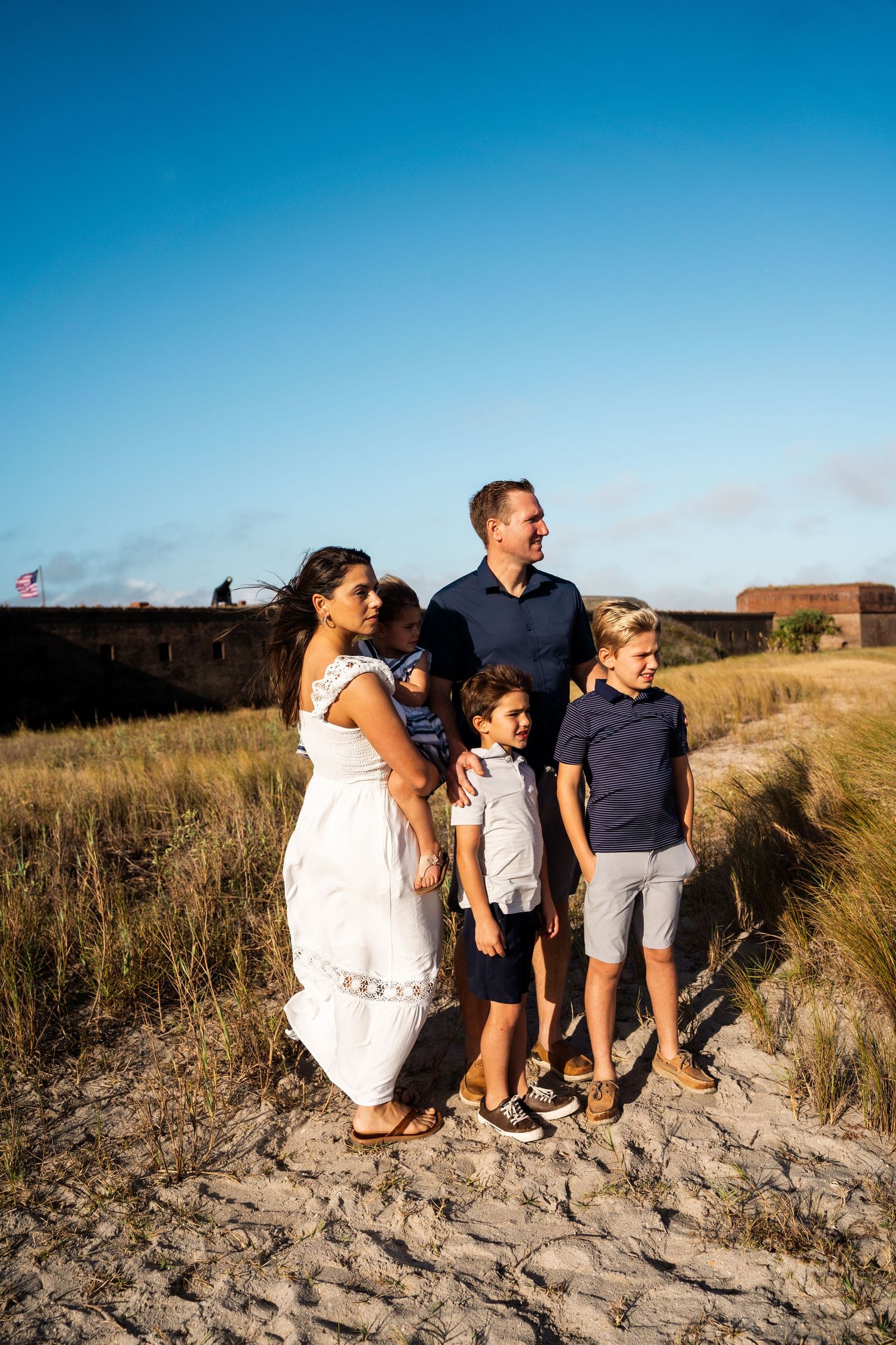 Family standing together on sandy path with blue sky and fortress wall in background.