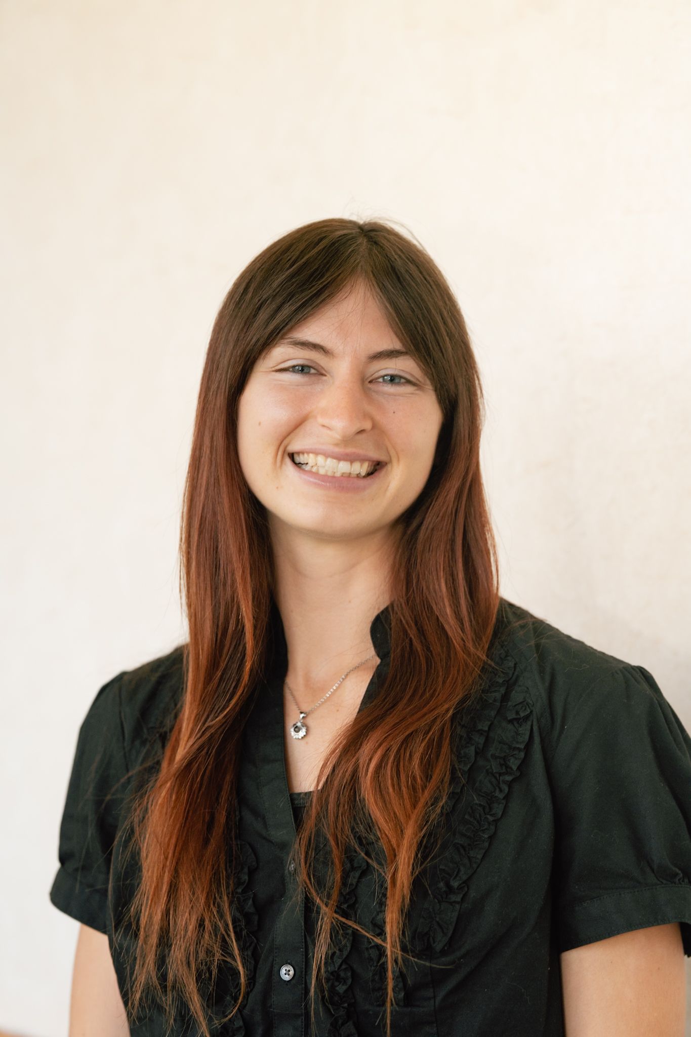Smiling young woman with long brown hair wearing a black blouse and necklace against a white background.