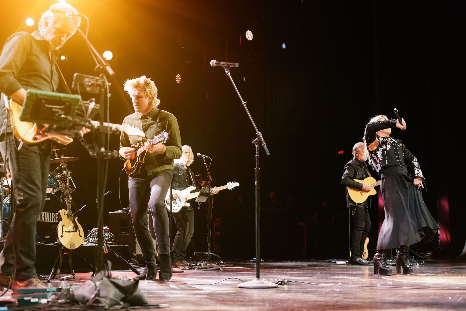 Live band performs on stage with guitarist in black hat and coat under warm spotlight at concert.