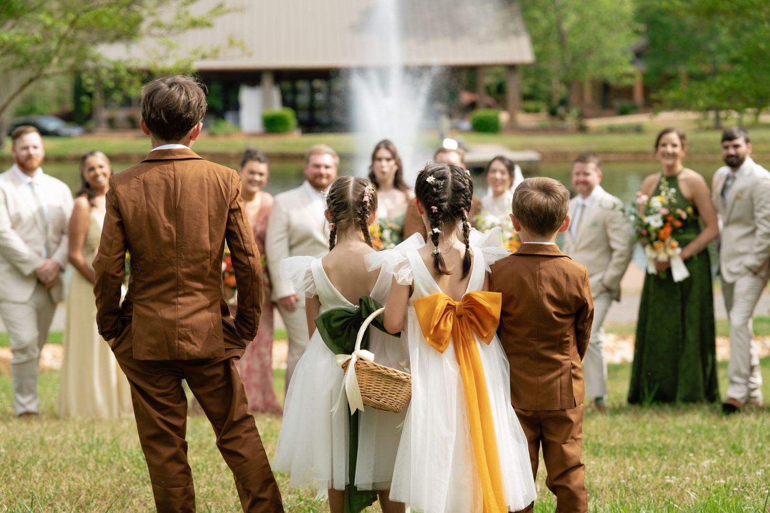 Outdoor wedding ceremony with flower girls, ring bearer, and wedding party gathered on green lawn near fountain.