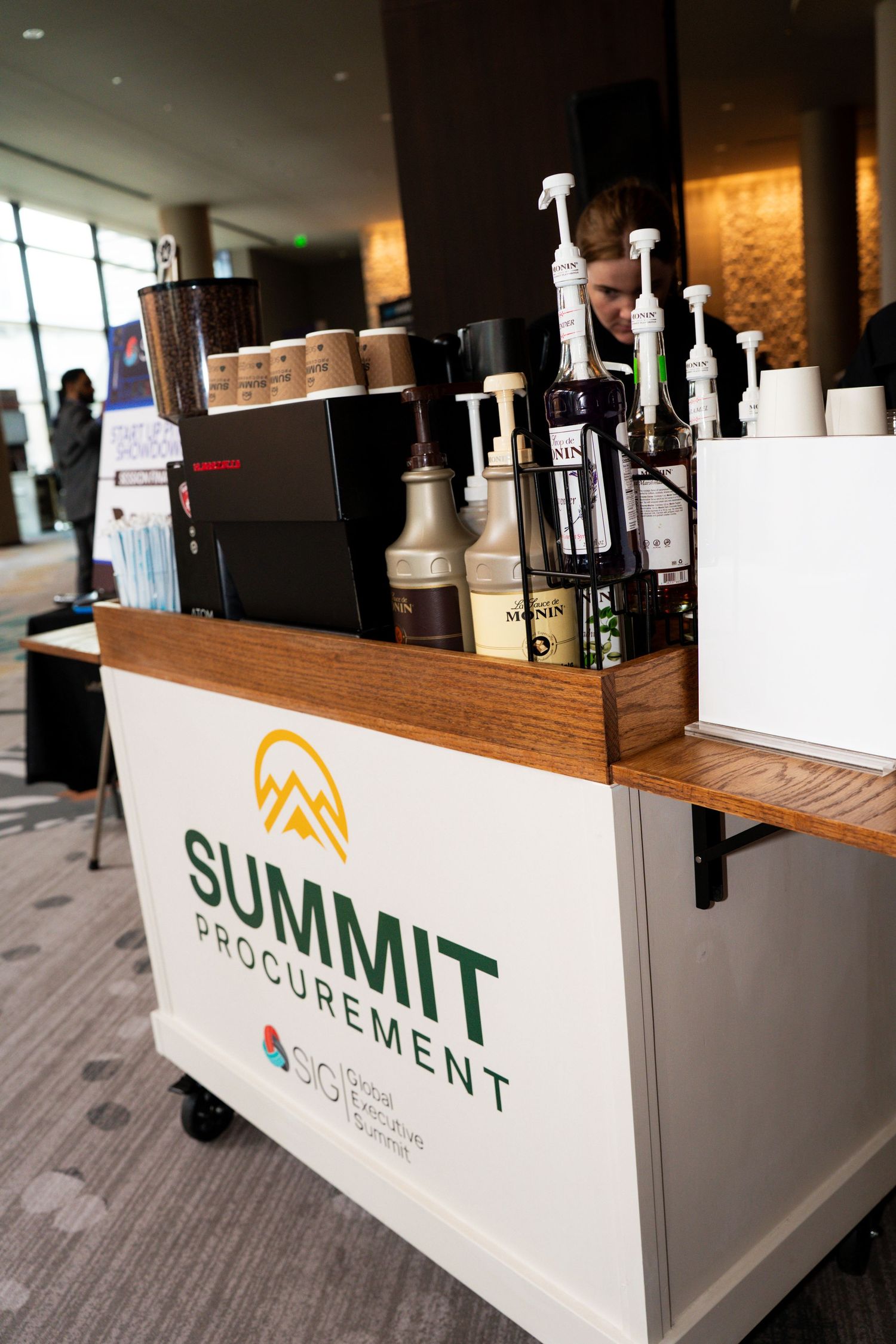 Summit Procurement branded coffee cart with Monin syrups and espresso machine at a corporate event.