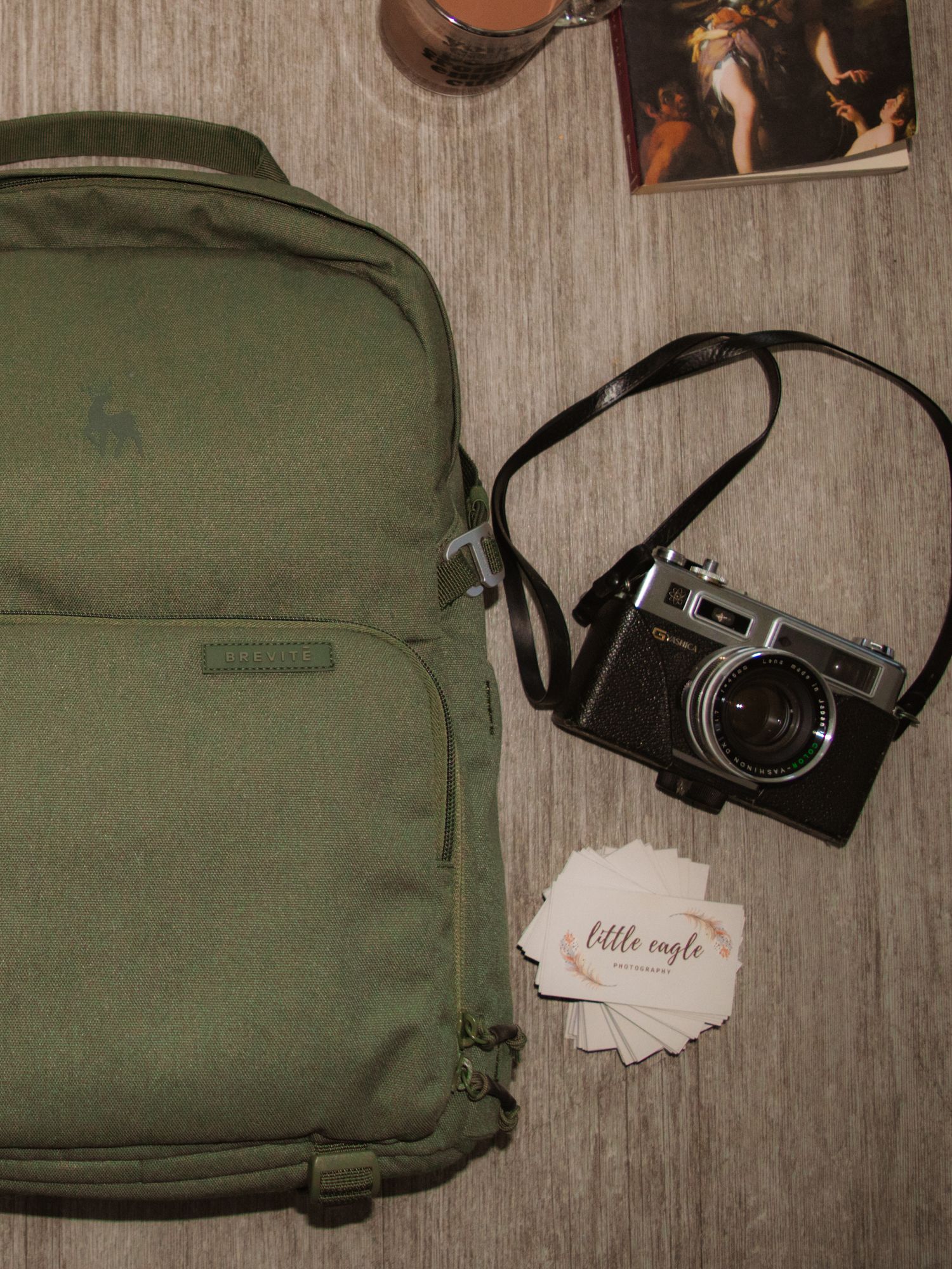 Vintage film camera placed next to an olive green backpack and scattered paper notes on a wooden surface.