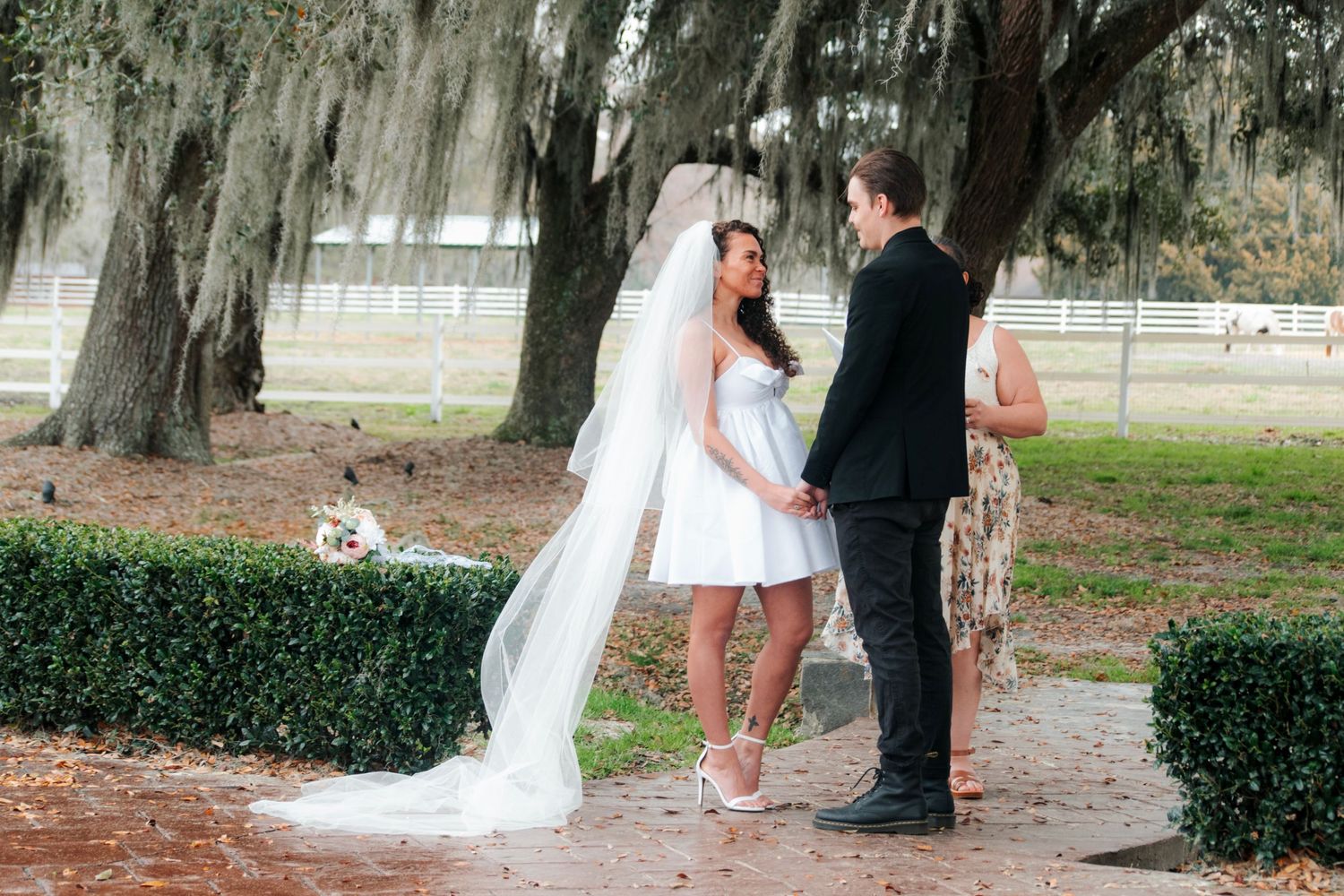 A couple in wedding attire stands beneath Spanish moss-draped trees in a romantic outdoor garden setting.