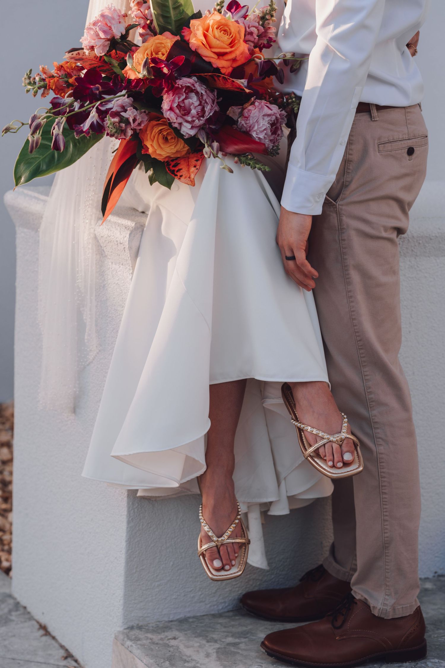 Elegant wedding photo showing white dress and khaki pants with vibrant orange and burgundy floral bouquet.