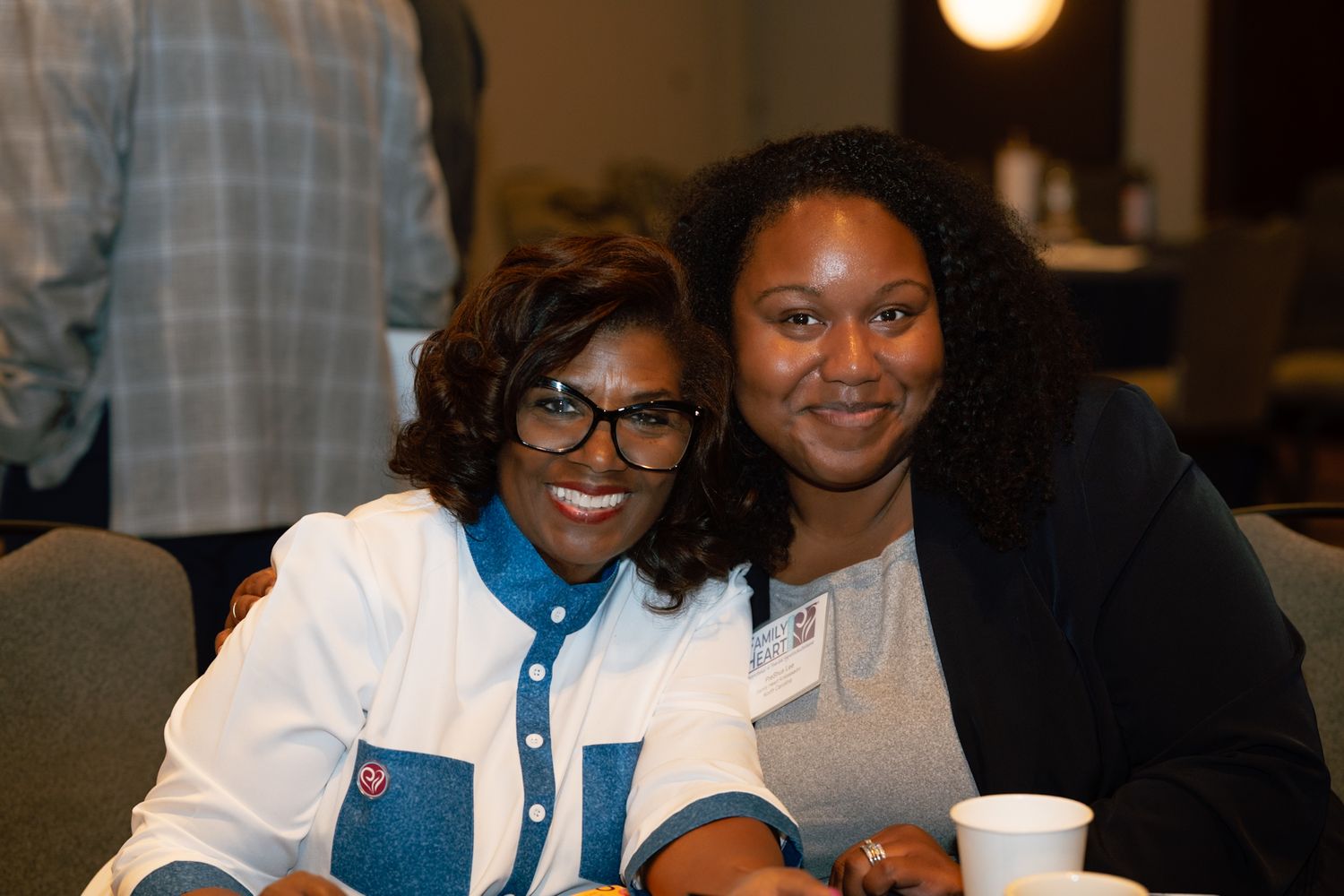Two women smiling and posing together at an indoor event, sitting at a table with a cup nearby.