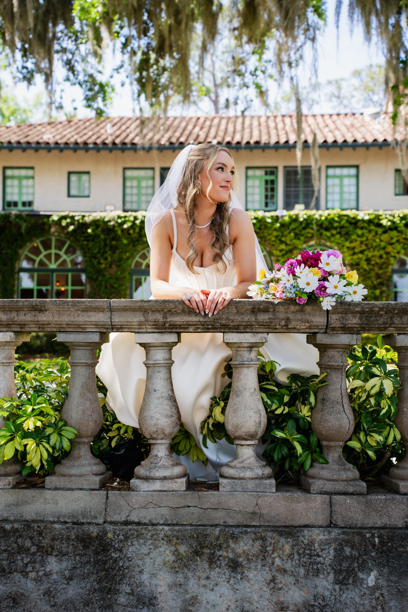 A bride in a white wedding dress sits on a decorative stone balustrade against a lush garden backdrop.