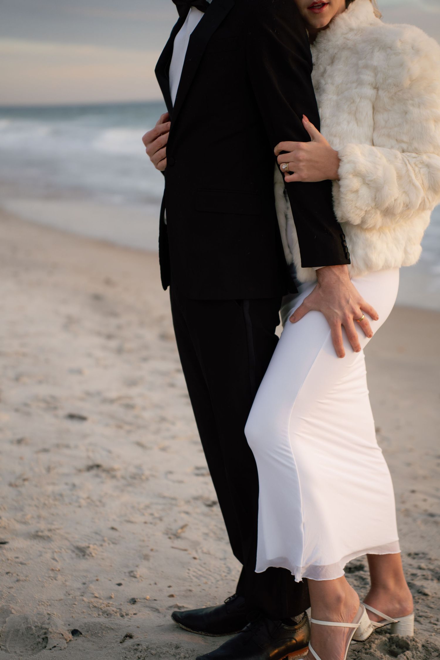 Close-up of couple's feet and legs walking together on beach with white dress and black suit details.