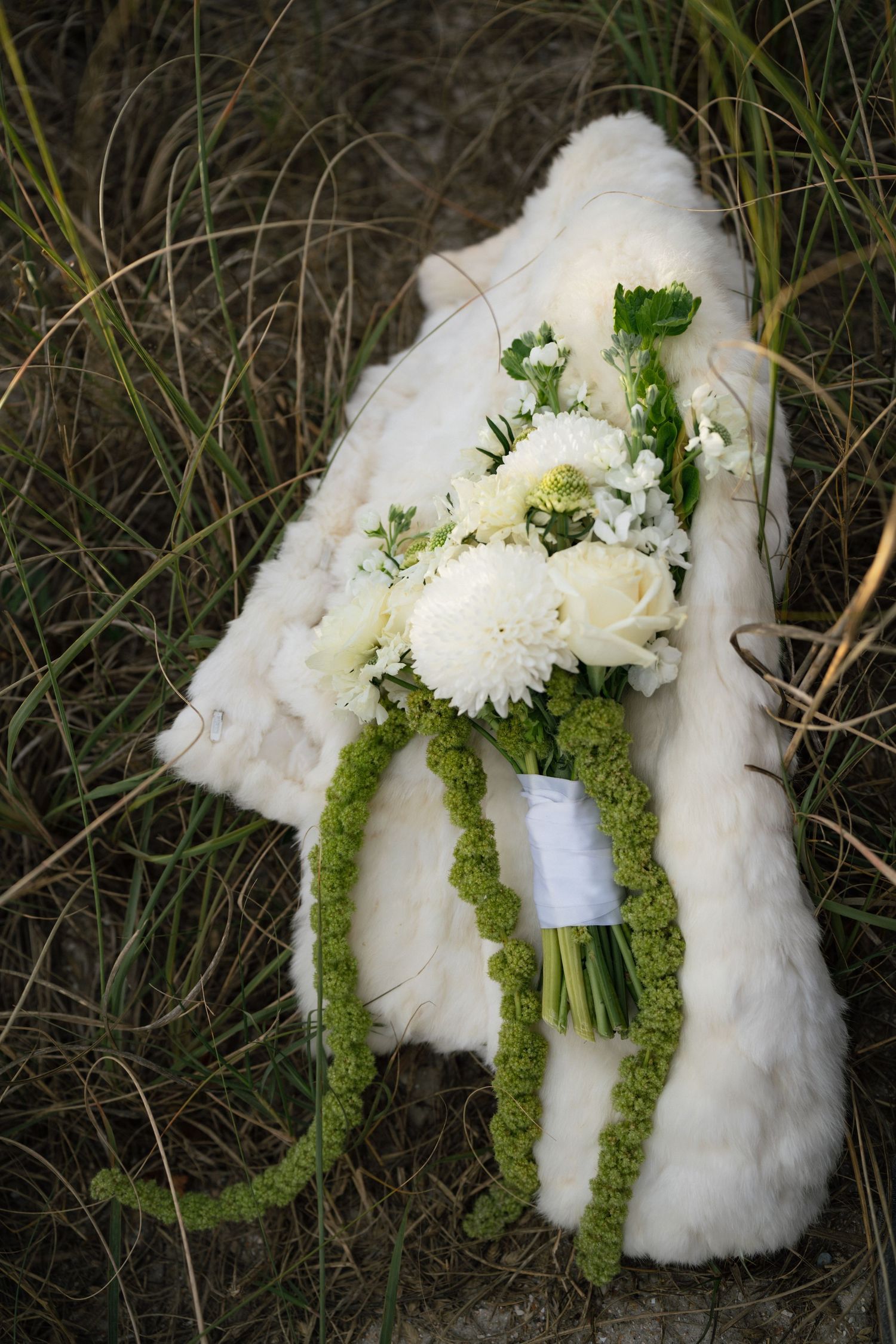 A white bridal bouquet with fresh flowers rests on a white fur wrap against grass background.
