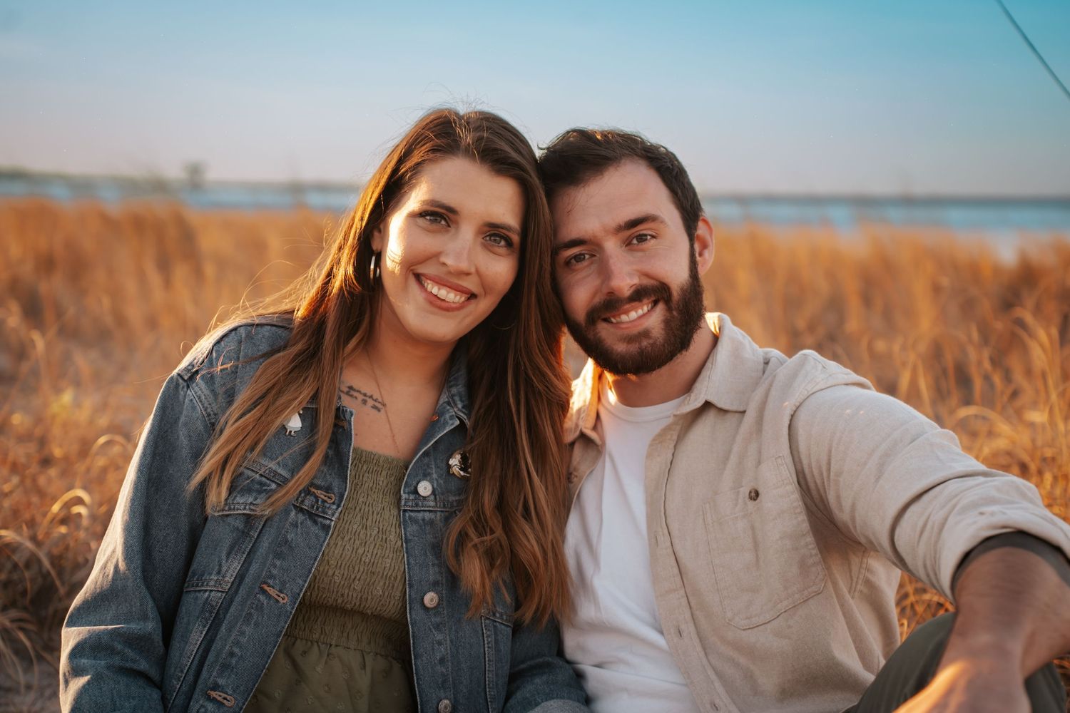 A couple smiles together in warm sunlight among tall golden grass near the water.