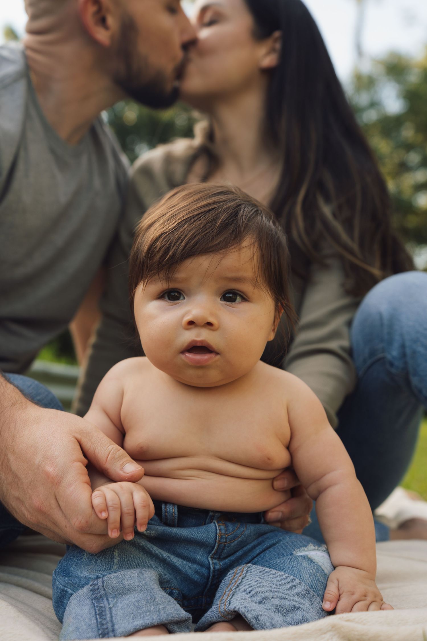 A chubby baby in jeans sits on a surface while parents embrace in the background.