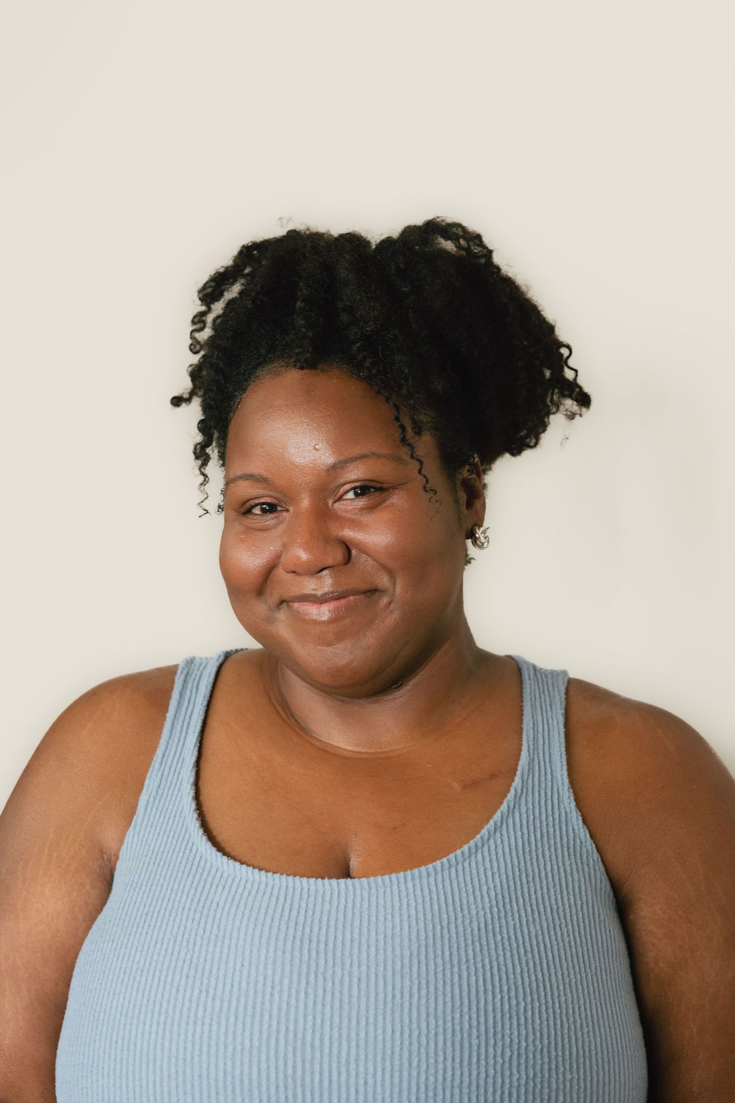 Smiling Black woman with natural curly updo hair wearing a light blue ribbed tank top against a neutral background.