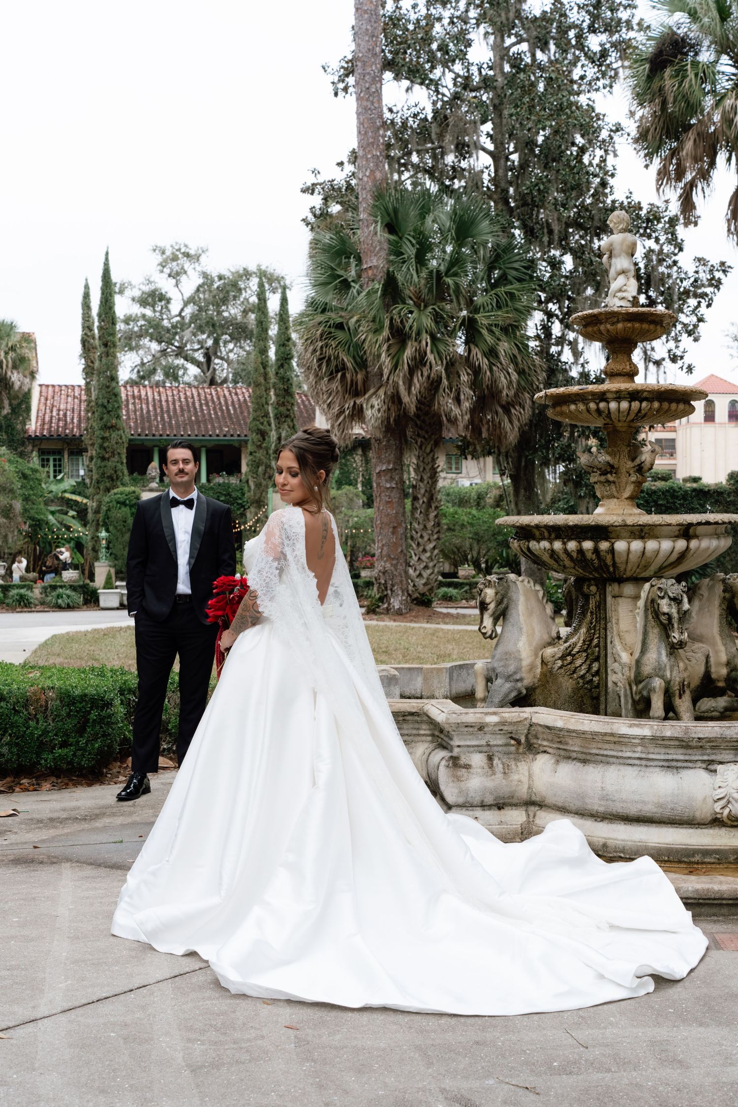 Wedding couple poses near an ornate stone fountain in a garden courtyard with palm trees and Mediterranean architecture.