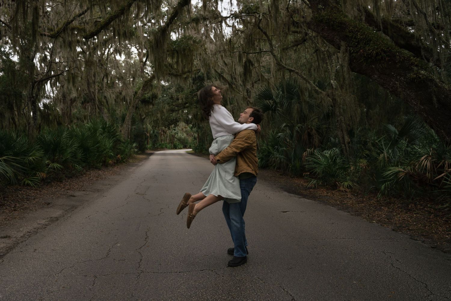 A couple embraces in a joyful lift on a tree-lined path covered with Spanish moss.