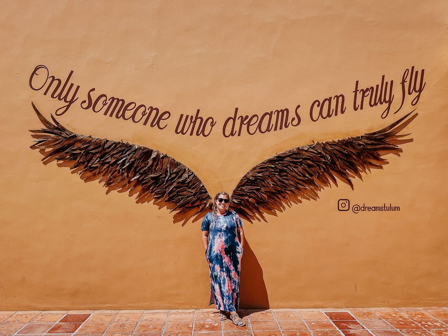 Street art mural shows large black wings against orange wall with inspirational quote about dreams and flying in Tulum