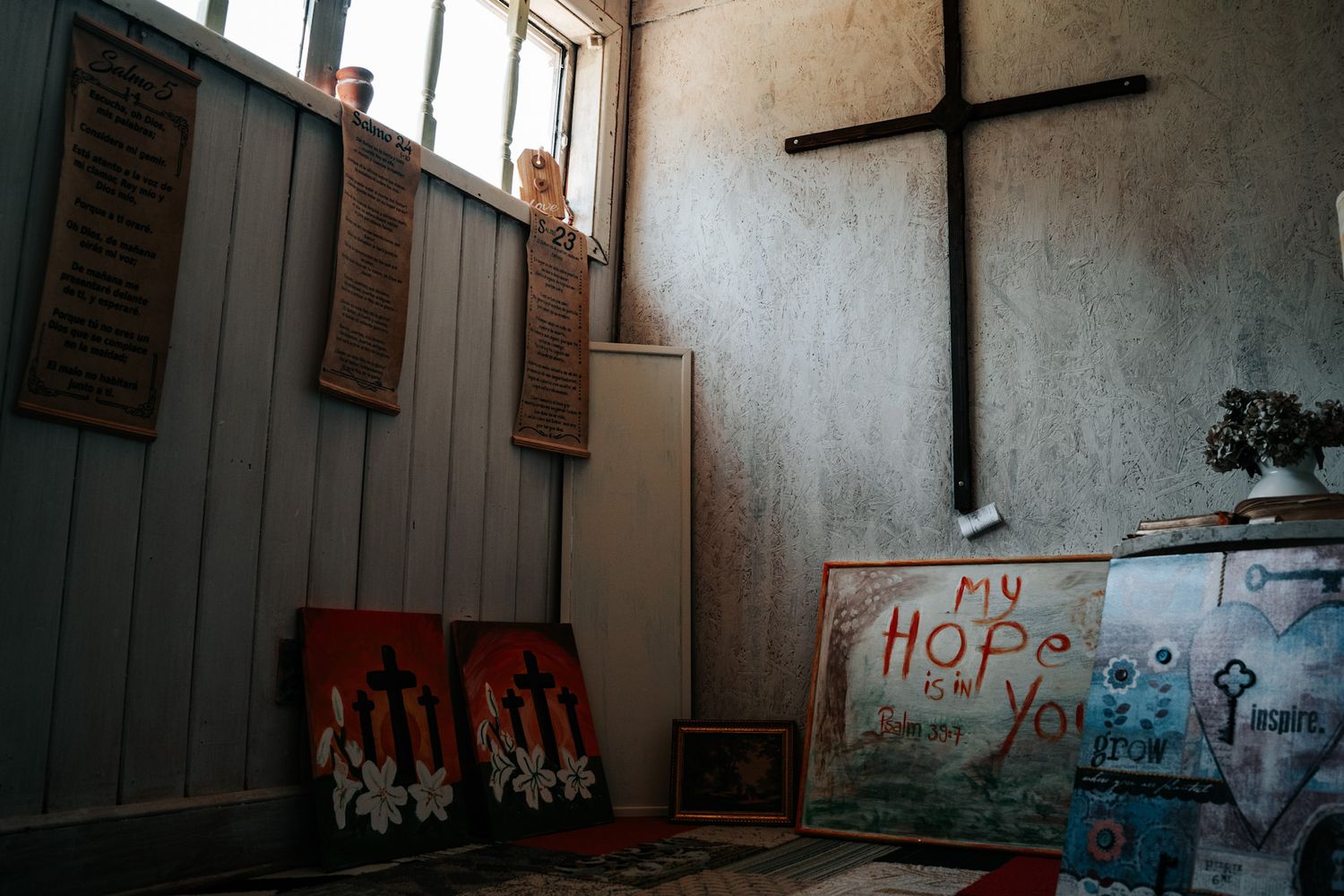A simple church interior with wooden shutters, a wooden cross on the wall, and graffiti art displayed on the floor.
