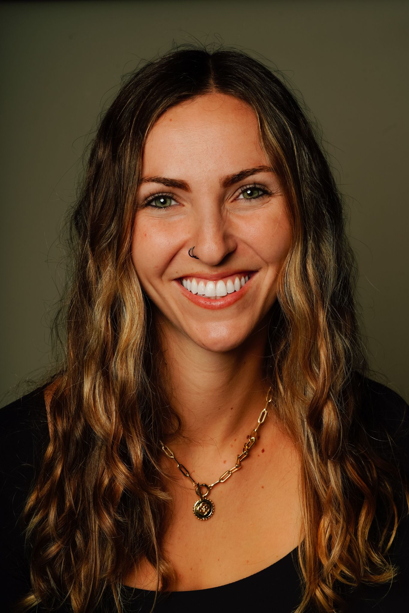 Smiling young woman with long wavy brunette hair, green eyes, and a gold chain necklace, wearing a black top.