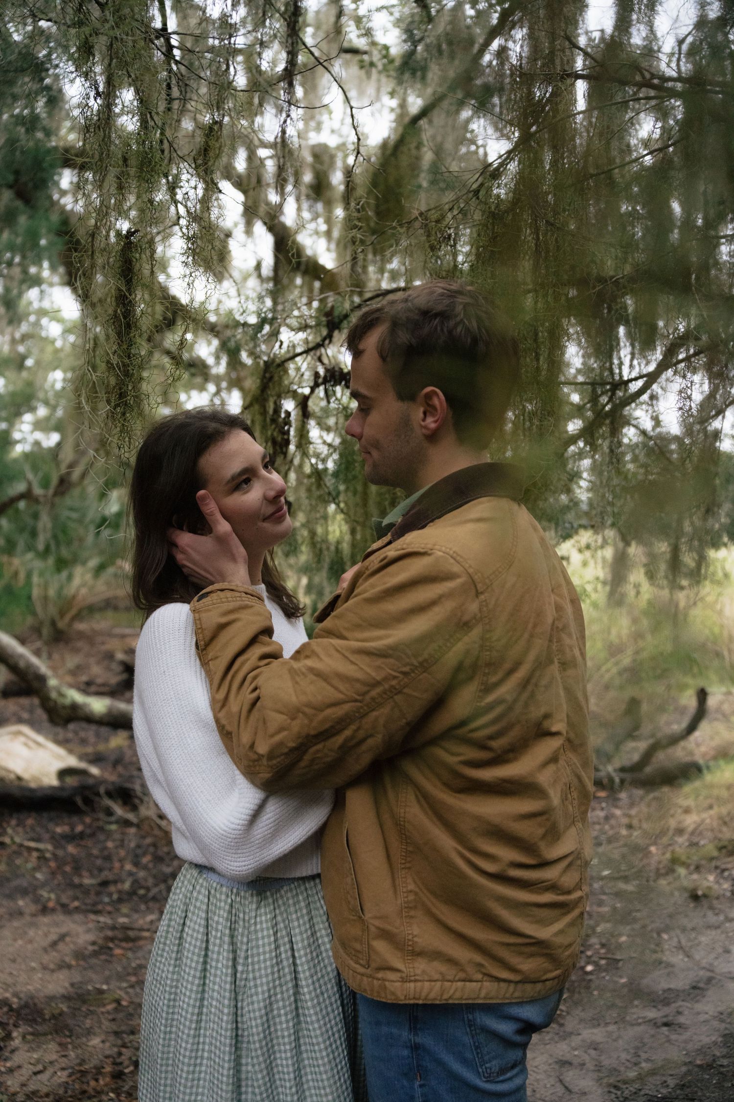A couple embraces in a romantic moment among tall trees in a wooded forest setting.