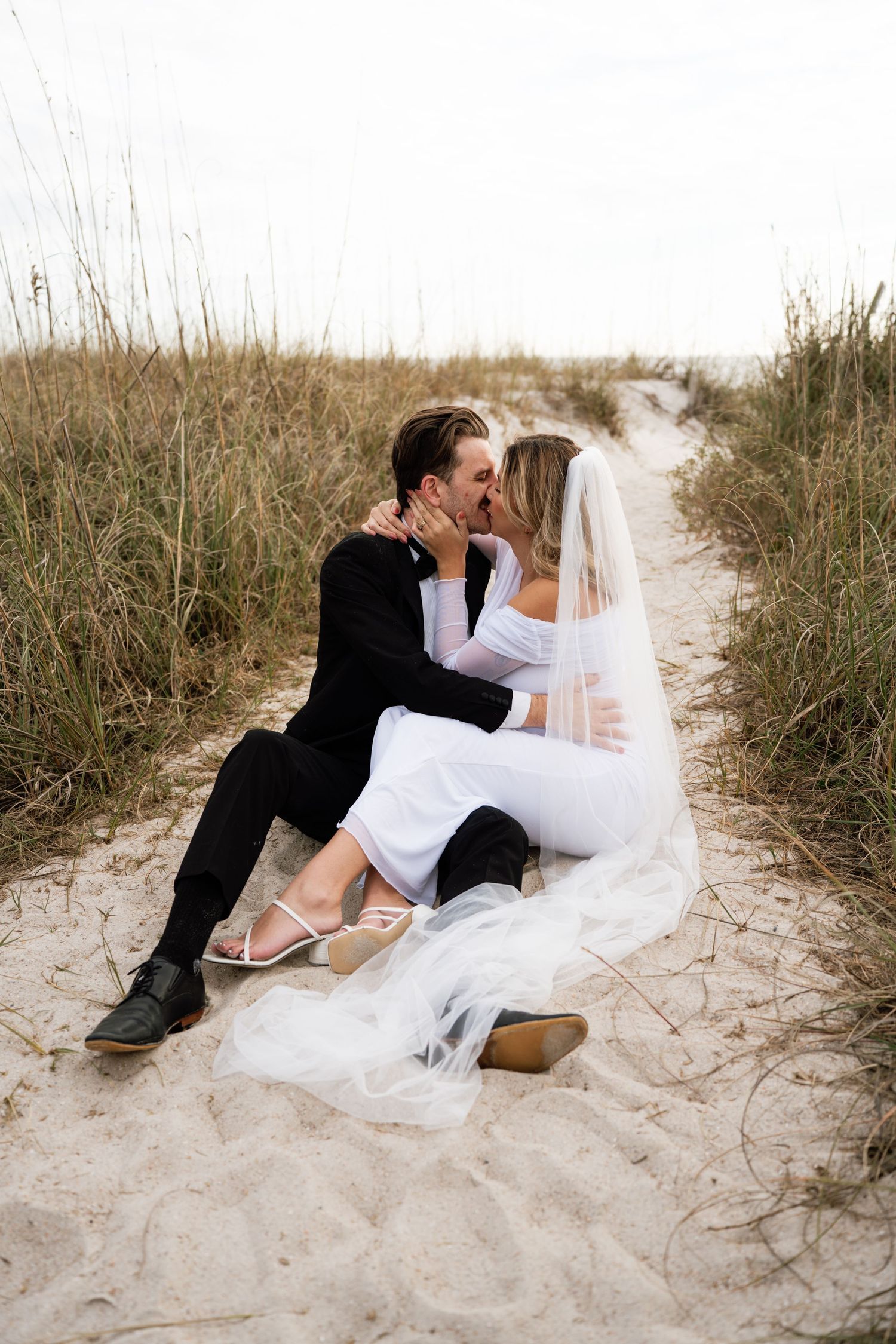 A romantic wedding couple shares intimate moments while sitting together on a sandy beach path.