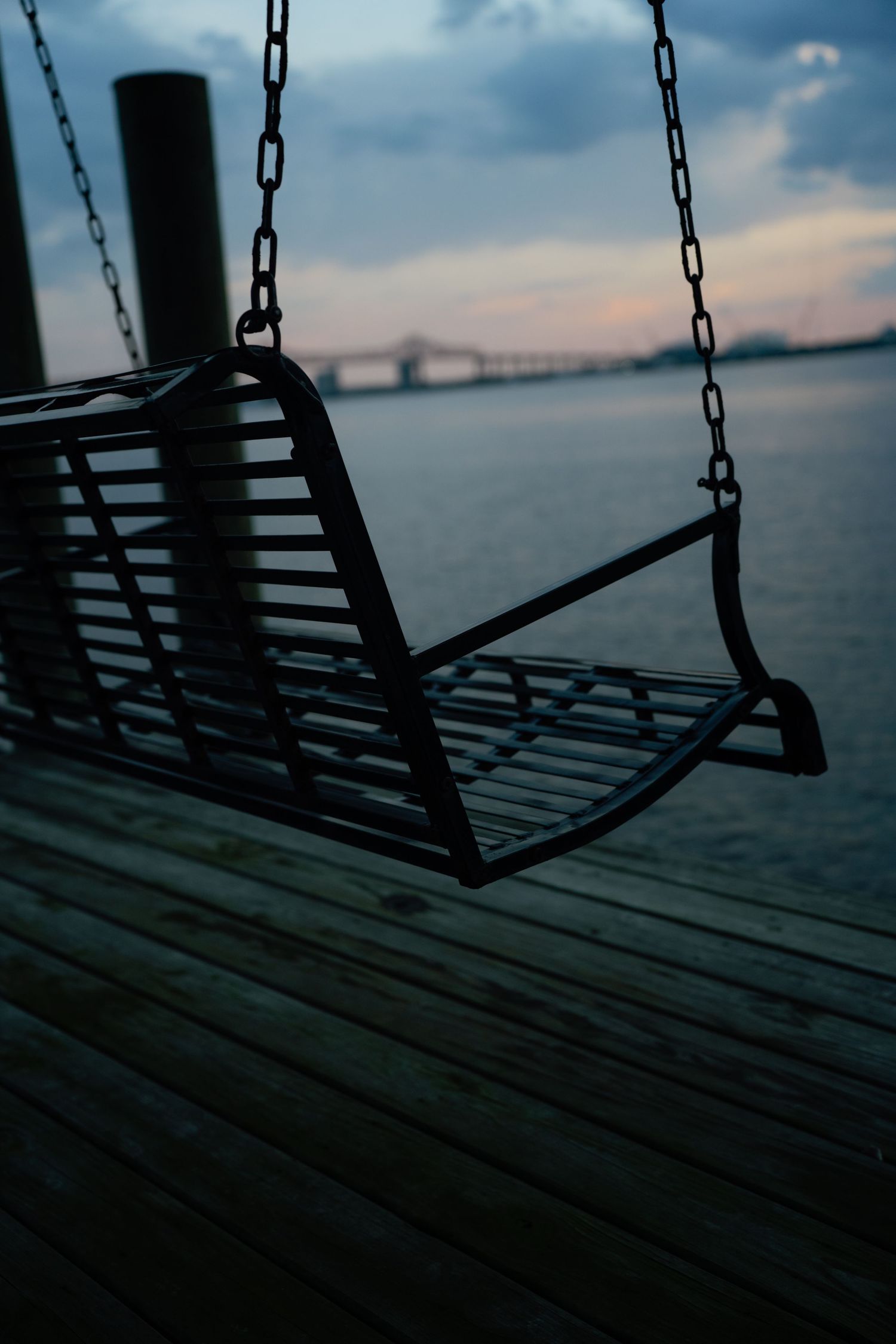 A silhouetted porch swing hanging by chains over a wooden dock at dusk with a calm waterway and bridge in the background.