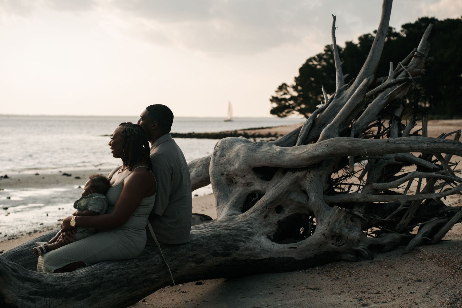 A couple sits on driftwood at the beach during sunset with a sailboat visible in the distance Fernandina Beach on Amelia Island, Florida