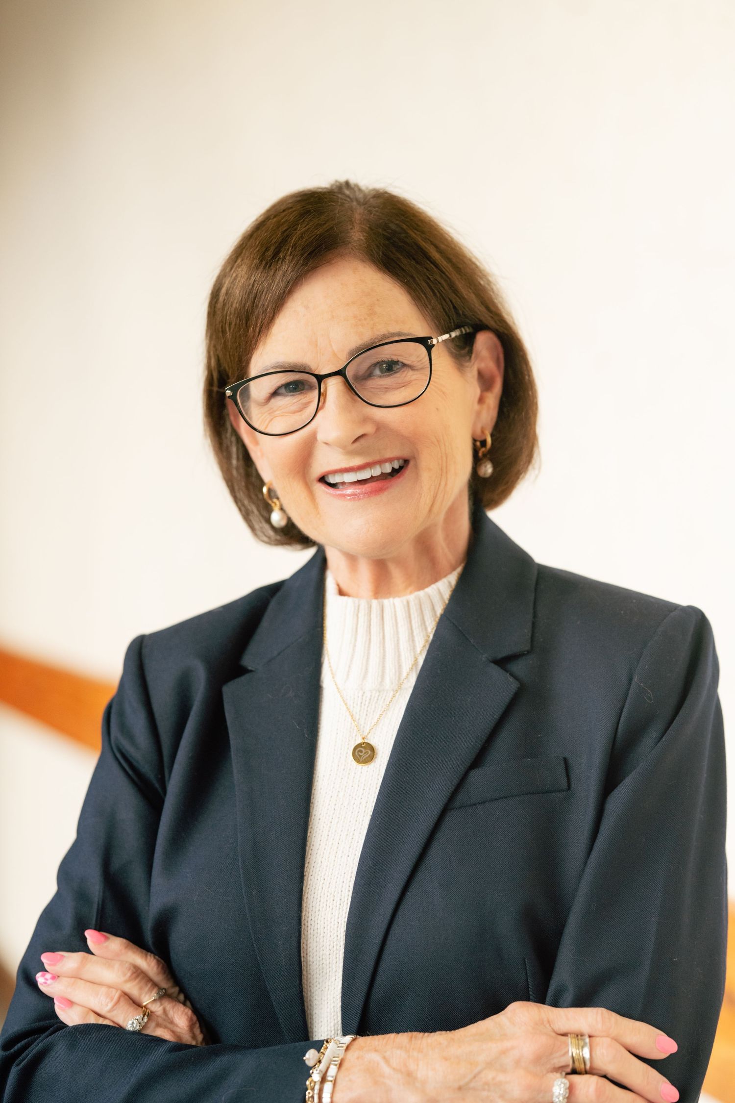 Professional headshot of a smiling senior woman in a navy blazer and glasses with arms crossed.