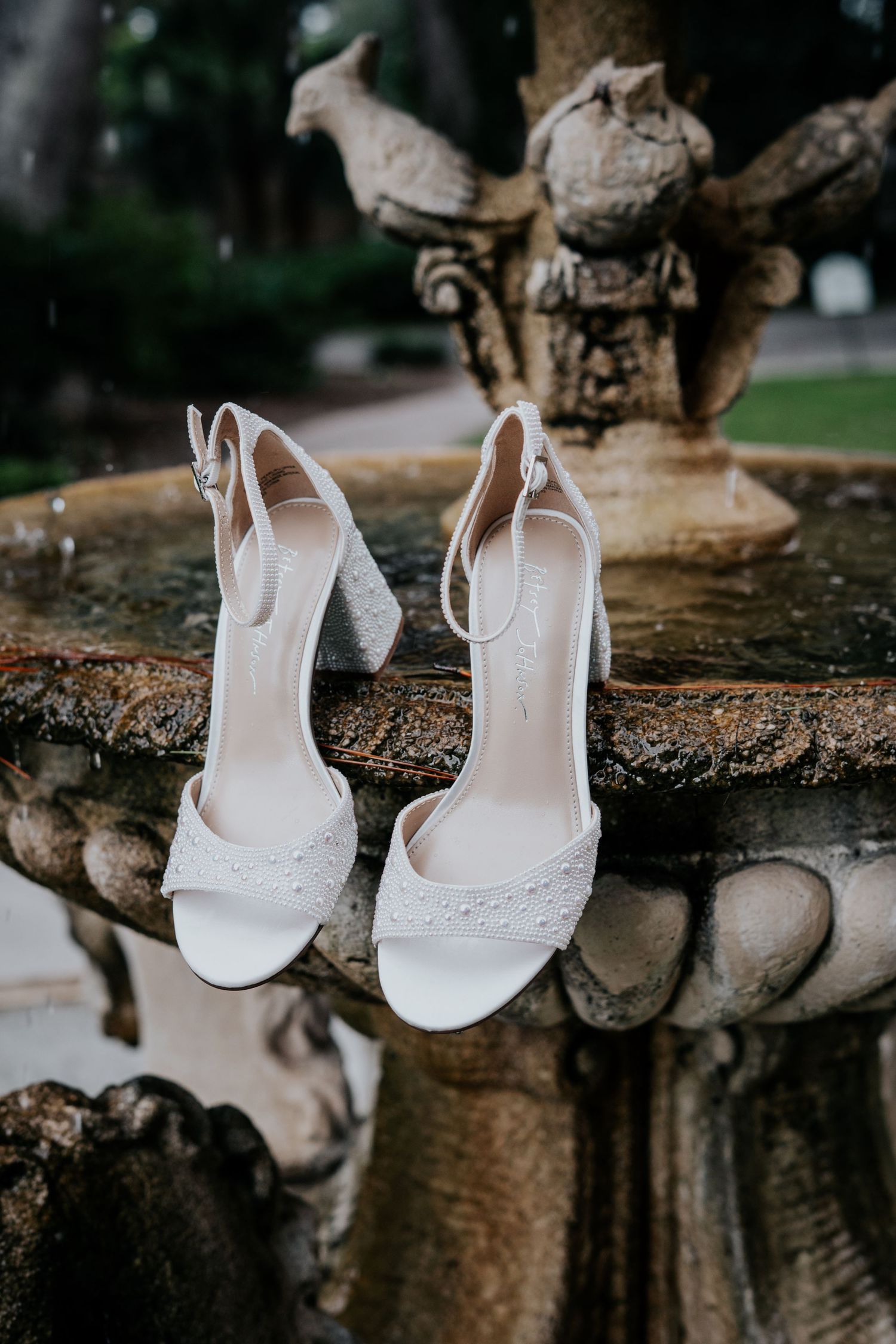 Elegant white bridal sandals with lace detail and block heels displayed on stone fountain ledge in garden setting.