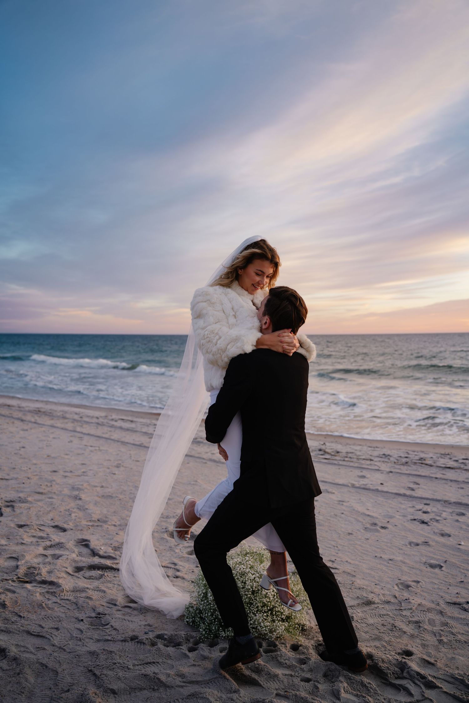 A couple in wedding attire dances playfully on a beach at sunset with ocean waves in the background.